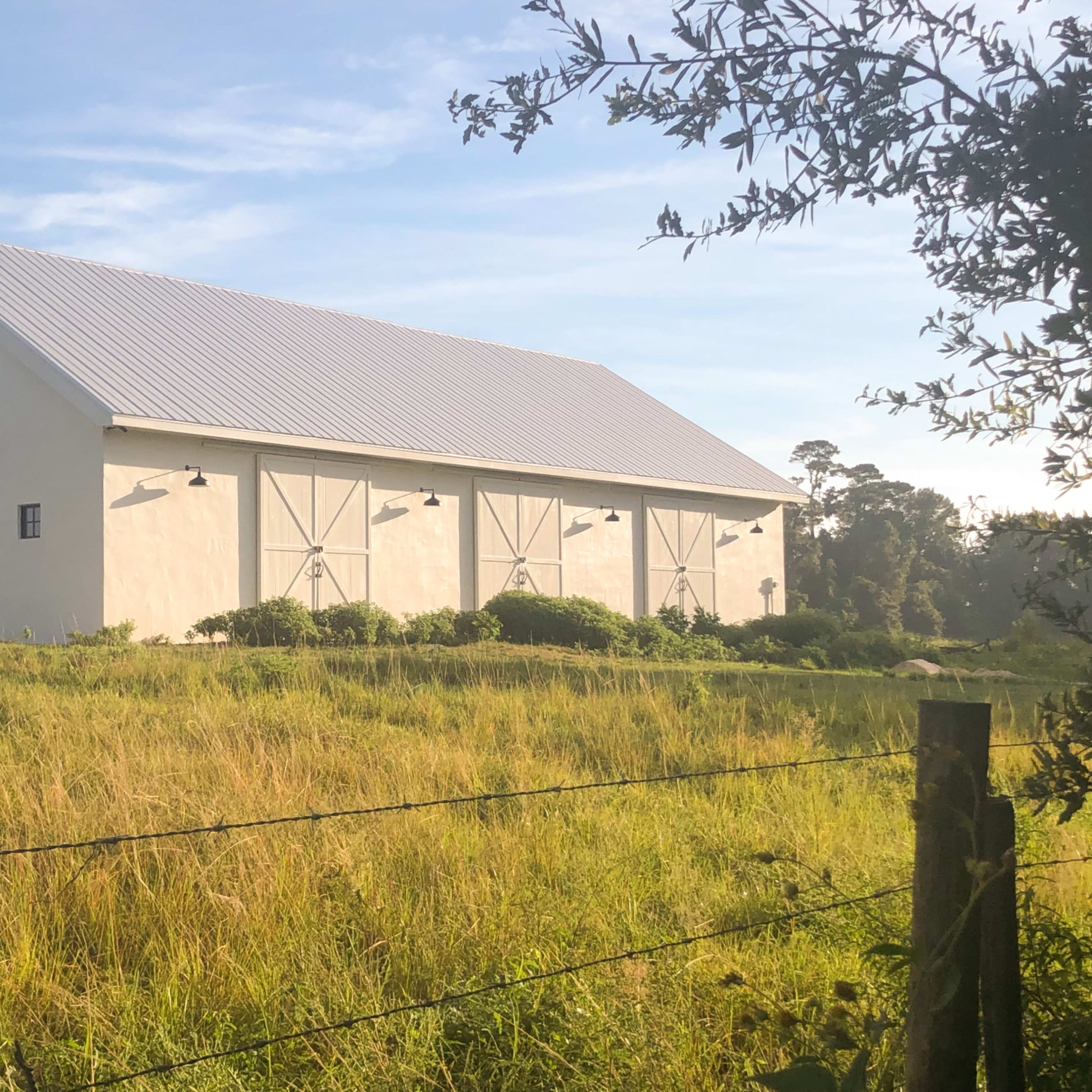 A large white barn with a metal roof sits in a grassy field, surrounded by trees.
