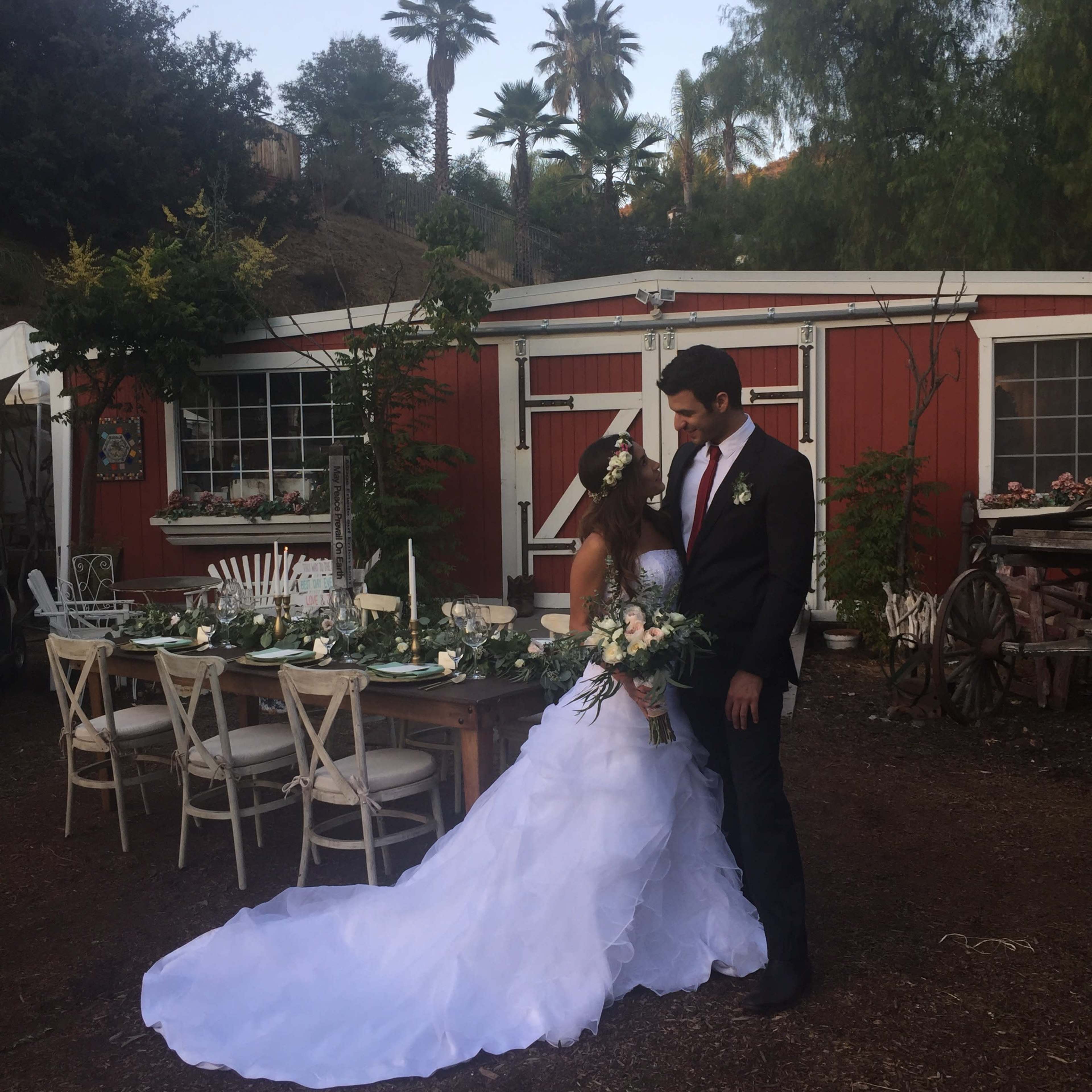 A bride in a white gown and a groom in a black suit stand together in front of a red barn, surrounded by greenery and a rustic table setting.