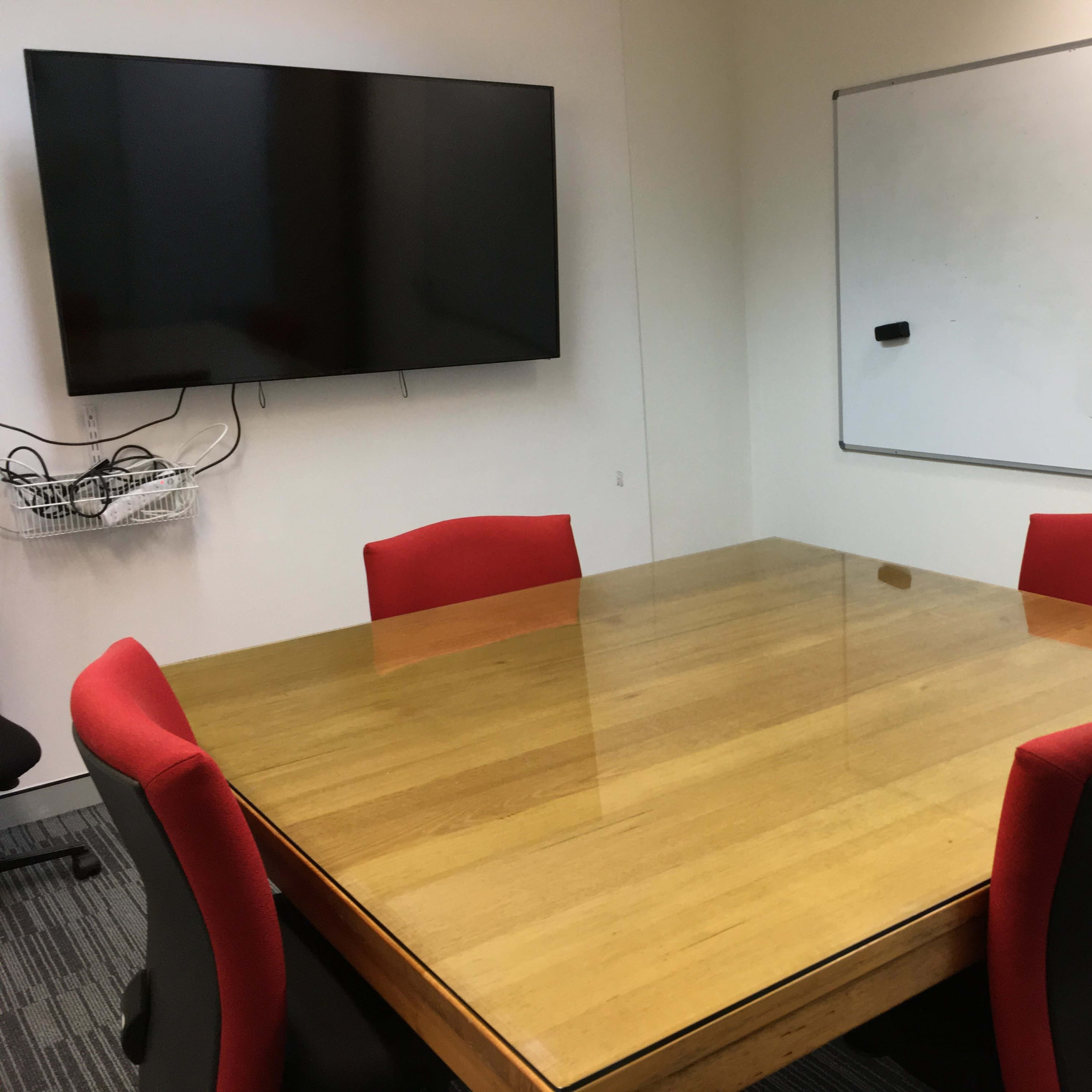 A conference room features a large wooden table surrounded by four red chairs, with a wall-mounted television and a whiteboard nearby.