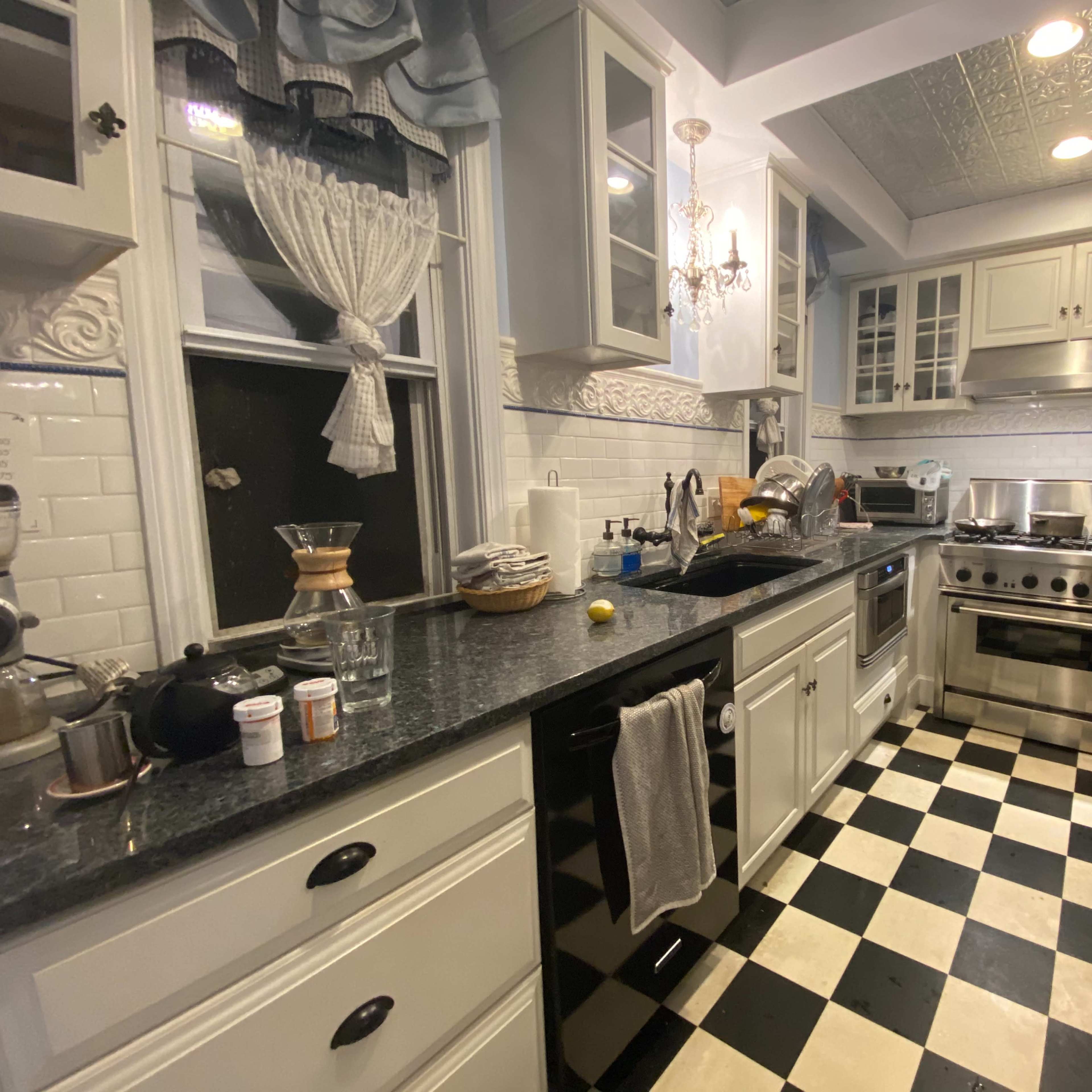 A kitchen with black and white checkered flooring, stainless steel appliances, and white cabinetry, featuring a mixture of kitchen utensils and clutter on the countertop.