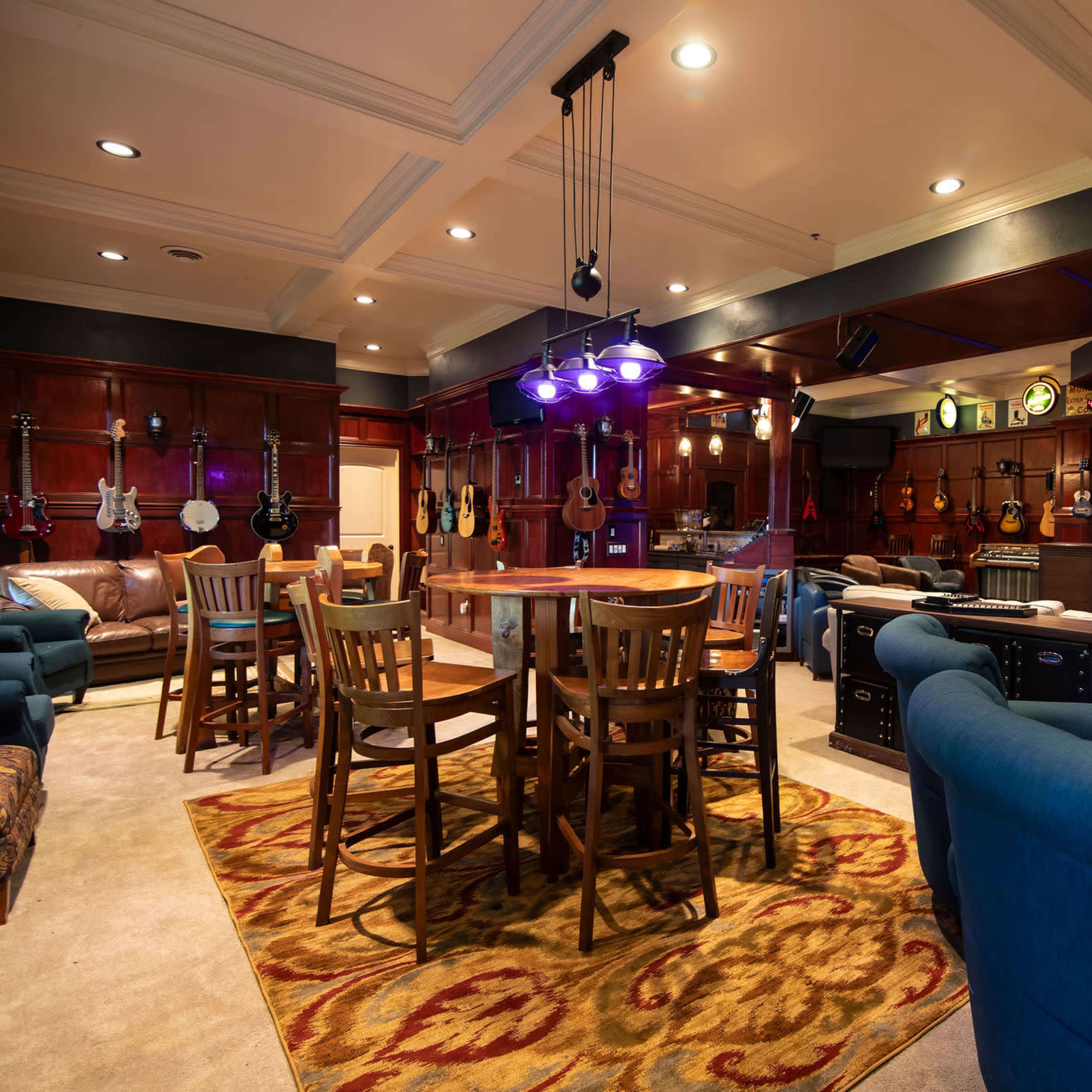 A cozy music room with wooden walls, various guitars displayed, seating areas, and a small stage illuminated in blue light.