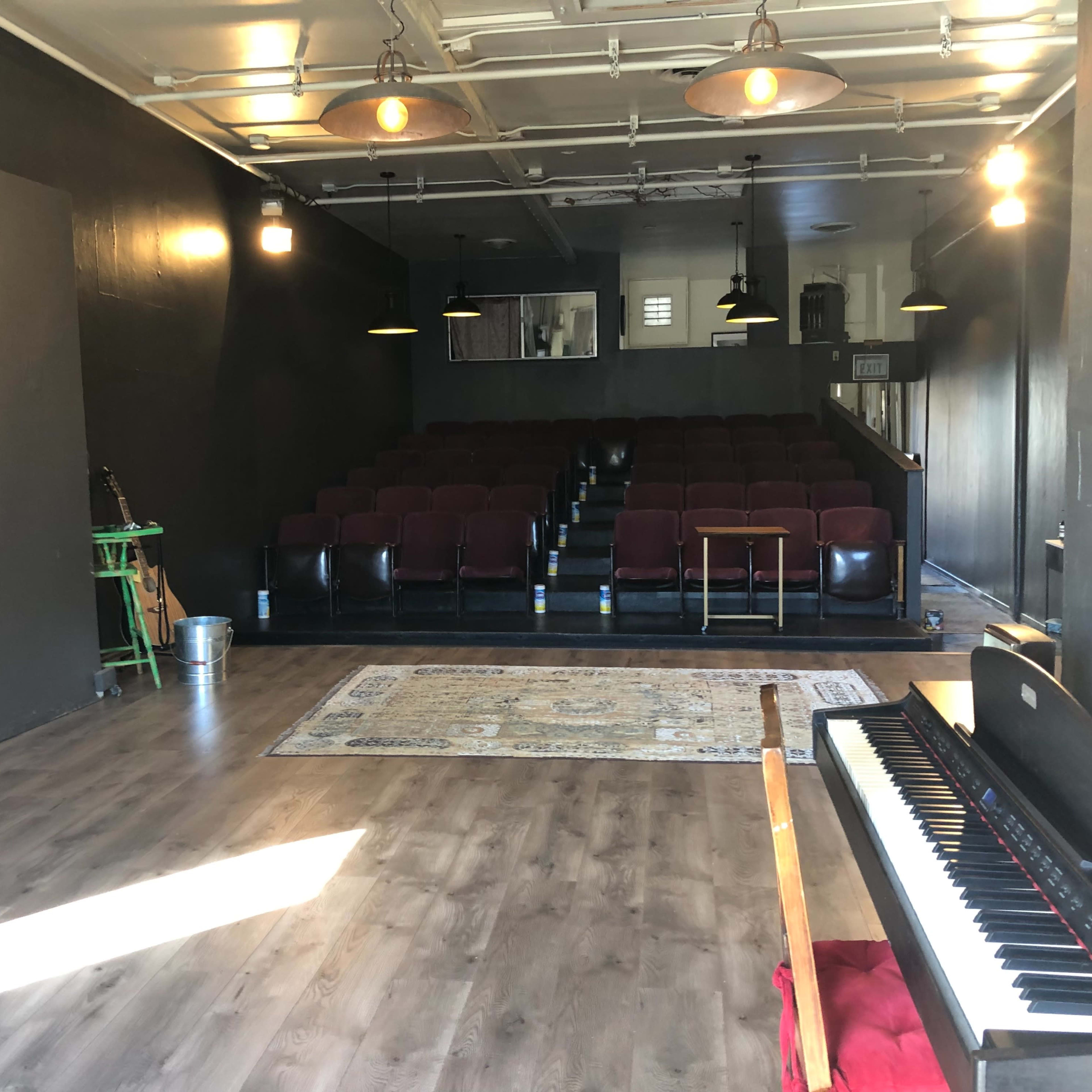 A performance space with a row of maroon seats, a piano in the foreground, and a patterned rug on a wooden floor.