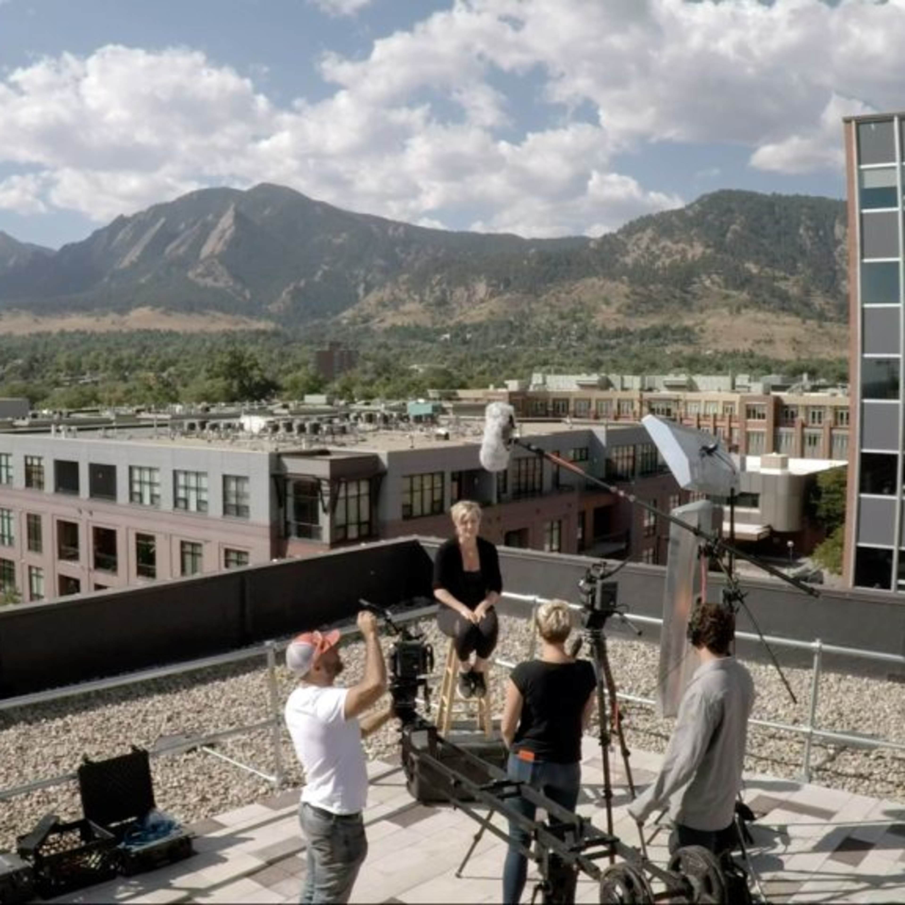 A film crew is capturing footage on a rooftop with a backdrop of mountains and urban buildings.