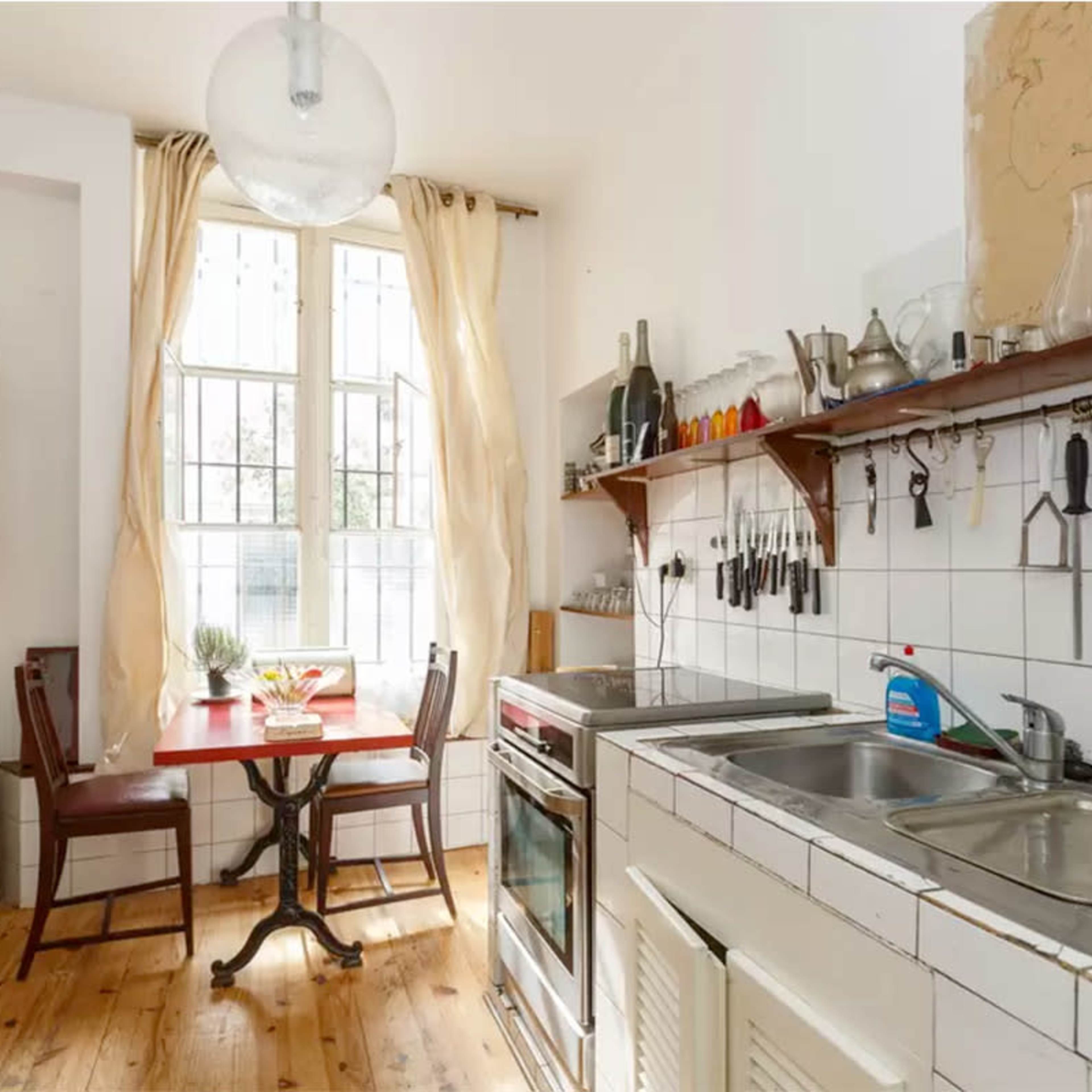 A compact kitchen features a wooden table with two chairs, white-tiled countertops, and shelves displaying glassware and utensils.