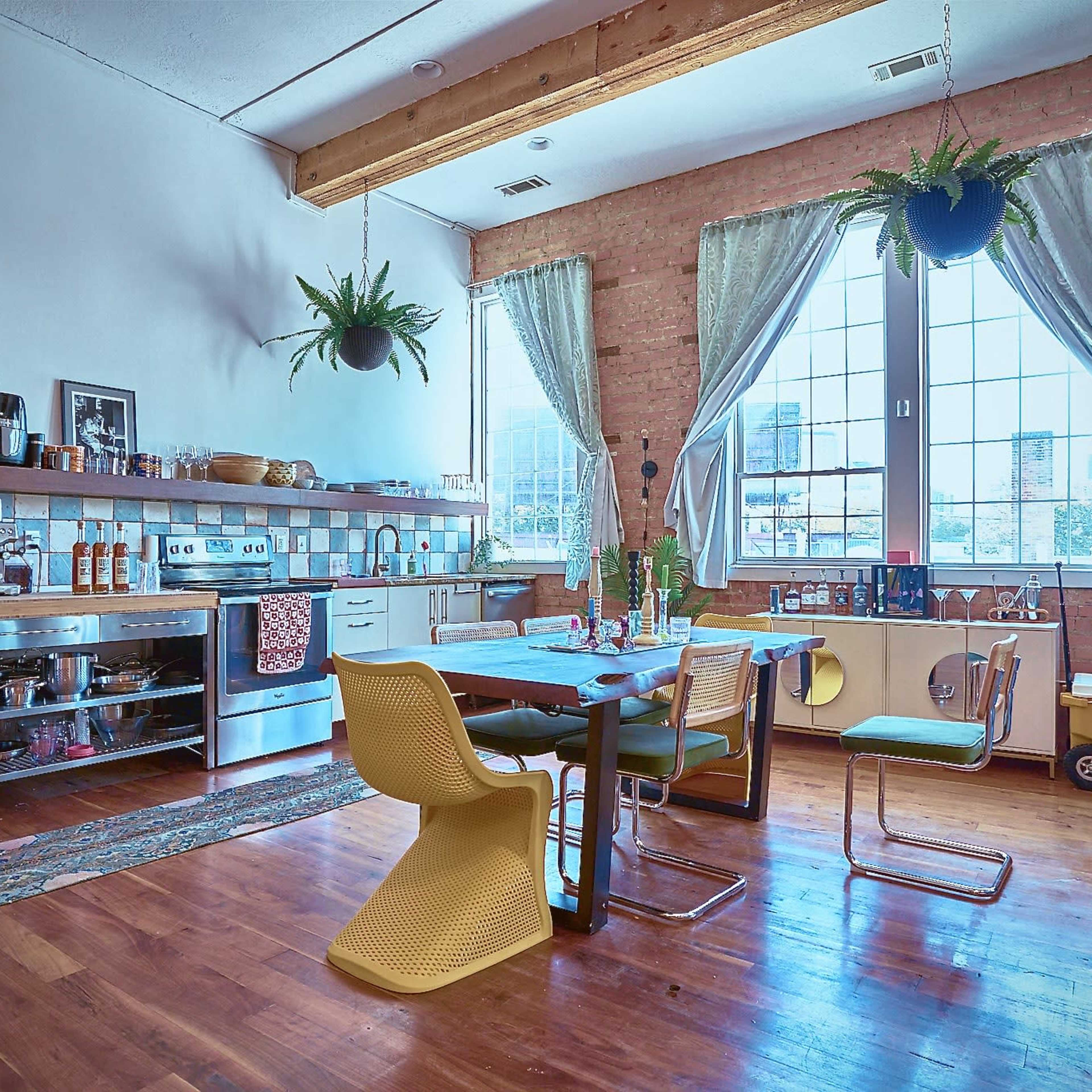 A spacious kitchen and dining area with wooden flooring, brick walls, a large window, and a combination of modern and vintage furniture.