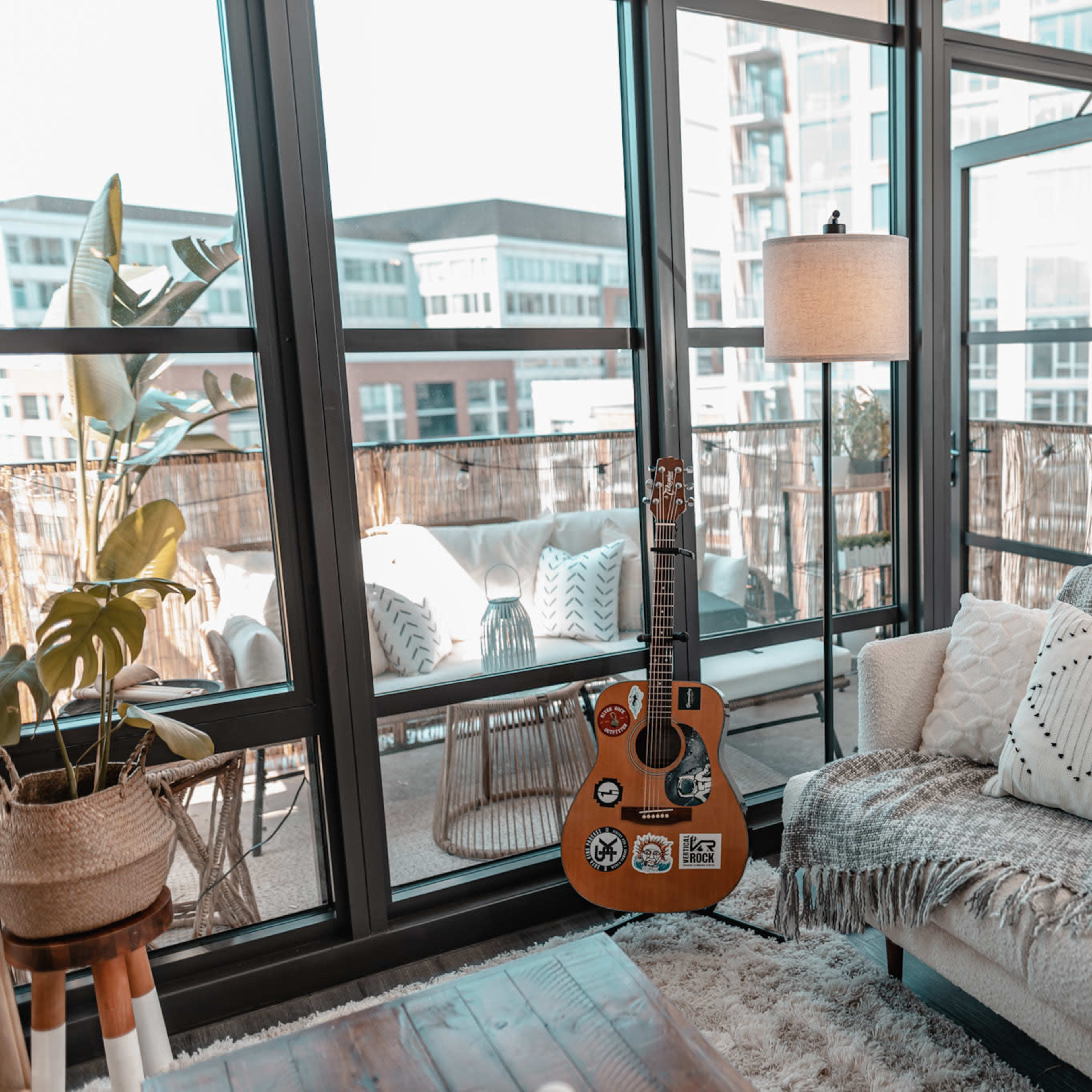 A guitar with stickers leans against a light-colored couch near large windows overlooking a balcony with outdoor seating.