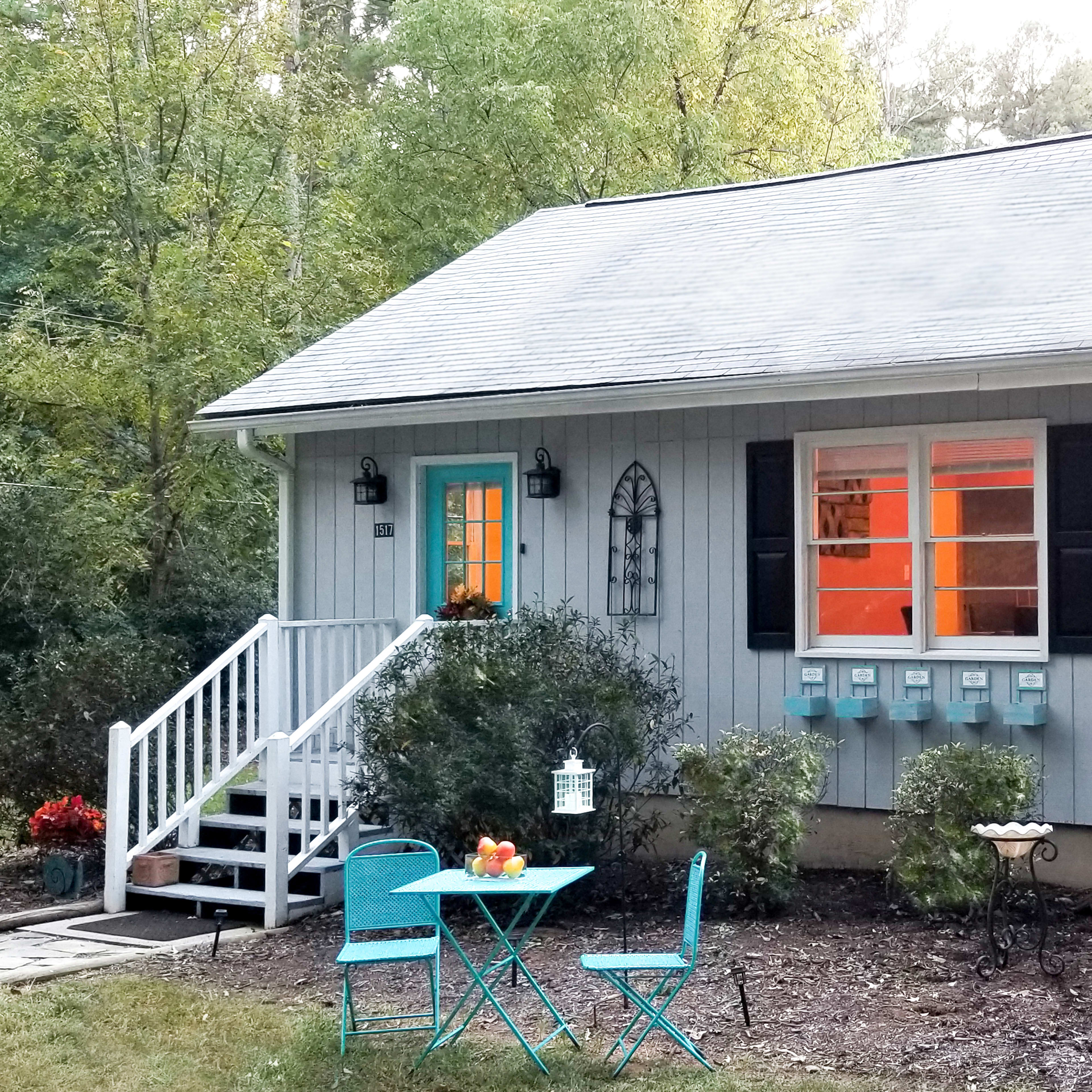 A small, gray house features a turquoise door and shutters, with a set of mailboxes and a blue table and chairs in the front yard.