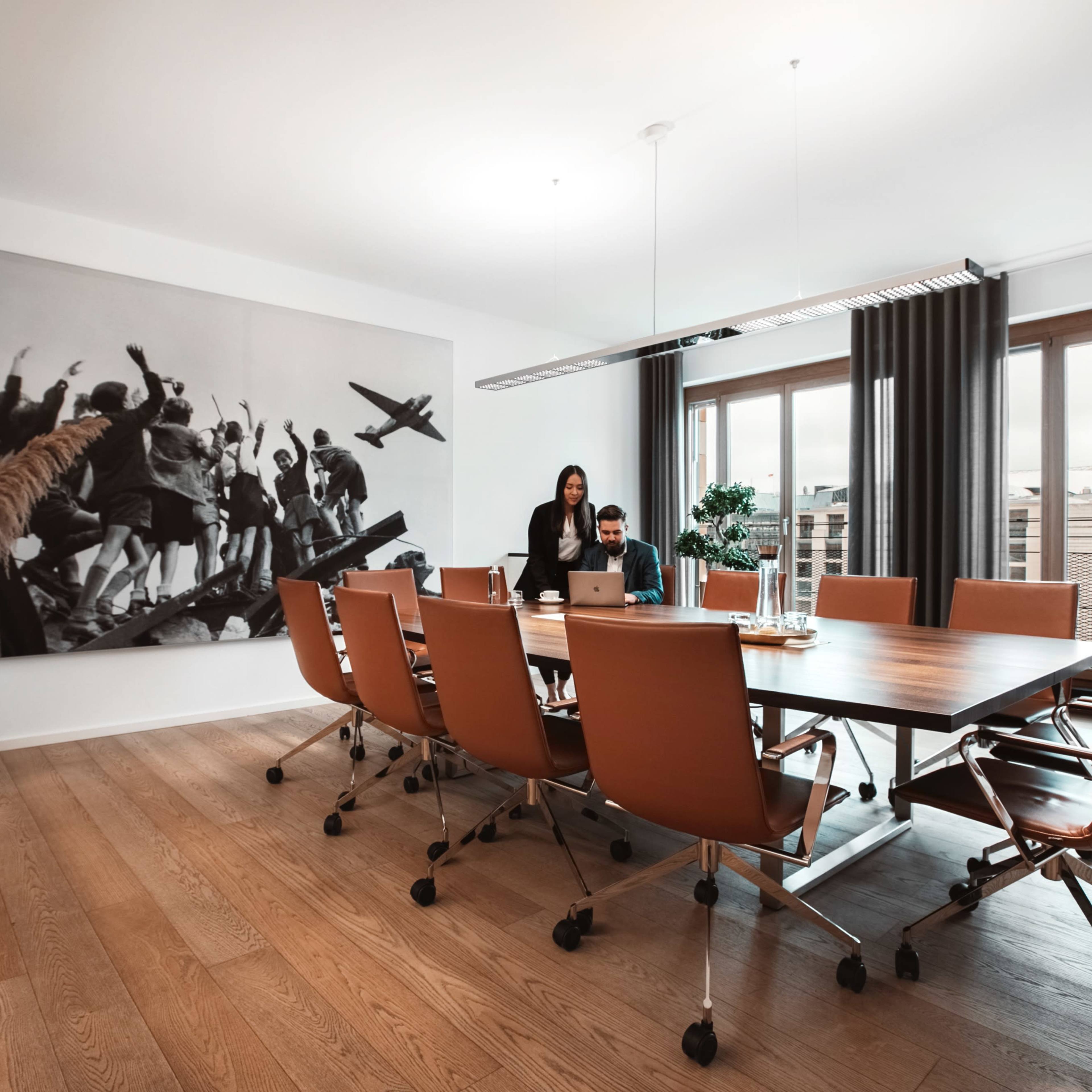 A modern conference room features a large wooden table surrounded by orange chairs, with a black-and-white mural on the wall and a group of people engaged in discussion.