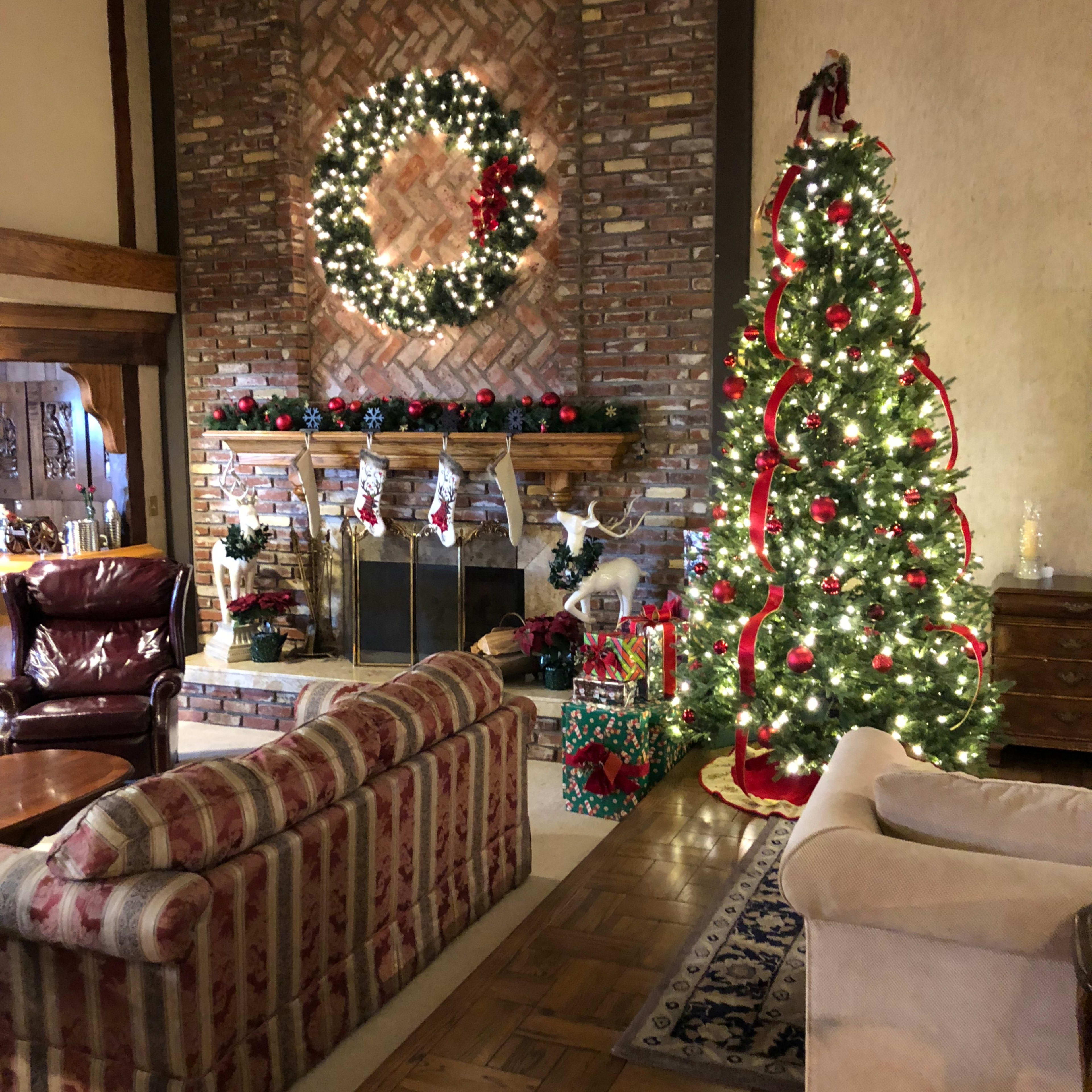 A warmly decorated living room features a stone fireplace adorned with a wreath and stockings, alongside a Christmas tree with lights and ornaments.