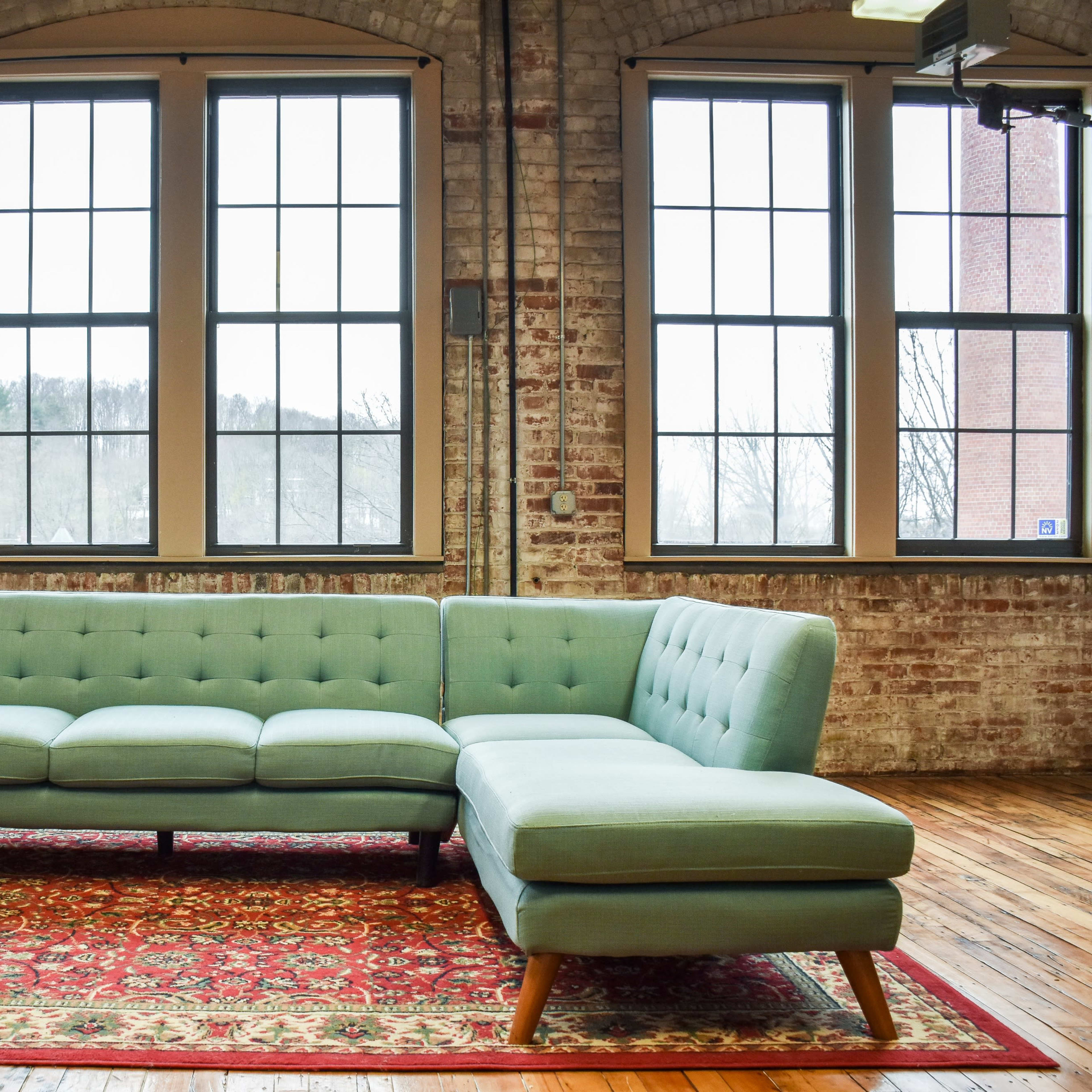 A light green sectional sofa is positioned on a patterned rug in a room with large brick walls and multiple windows.