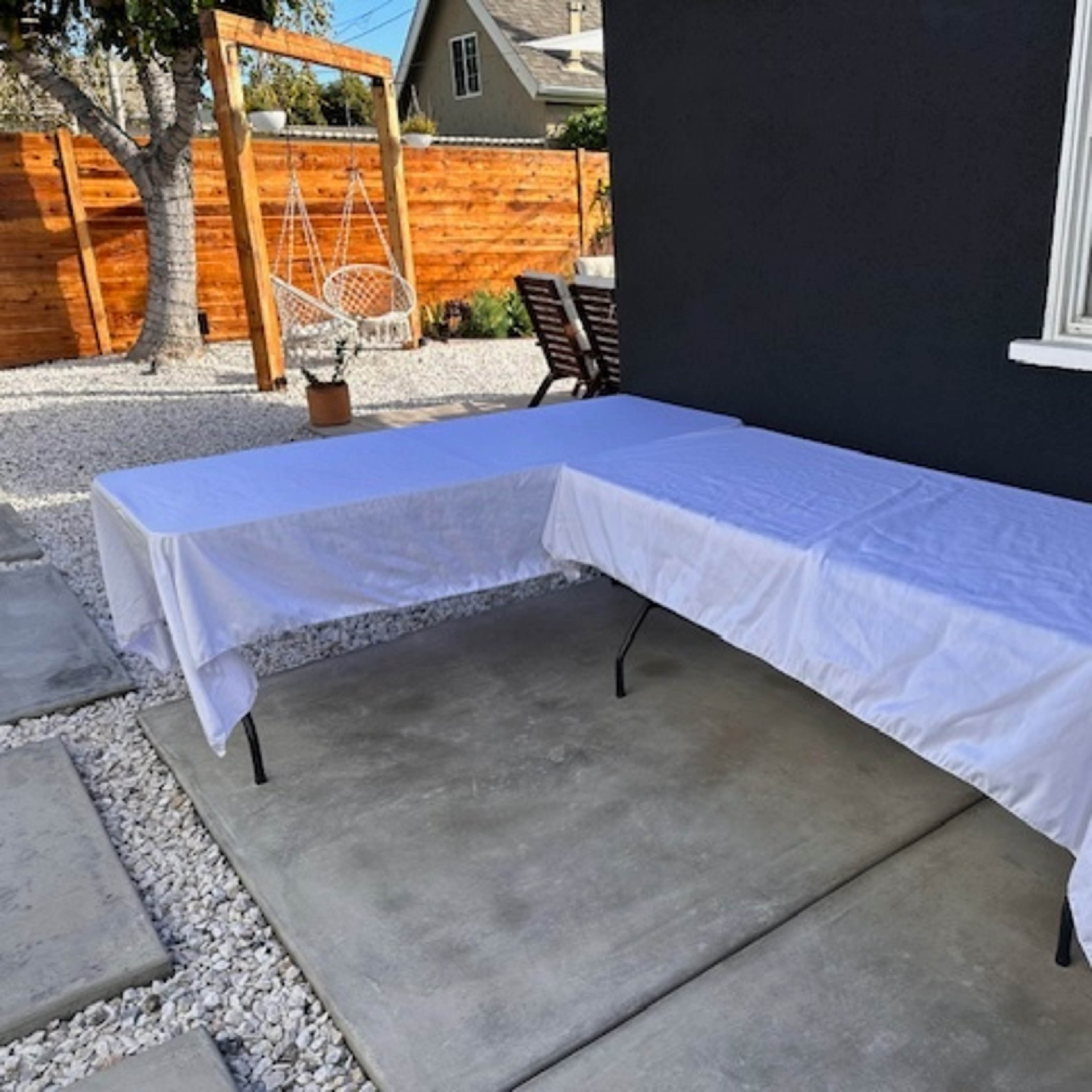 Two tables covered with white tablecloths are set on a concrete patio outside a house, with a garden area in the background.