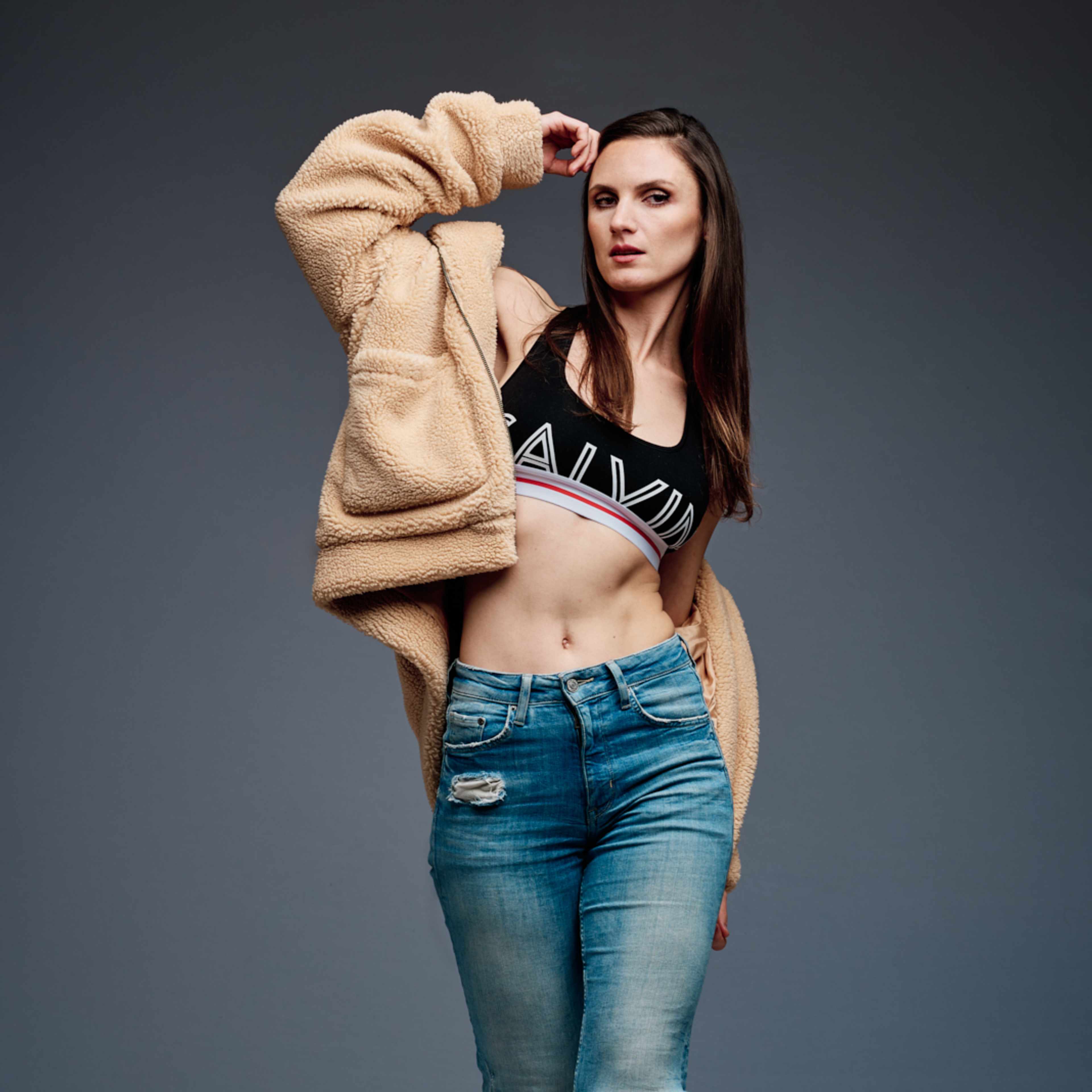 A model poses confidently in a cropped top and ripped jeans, wearing a fuzzy beige jacket against a gray background.