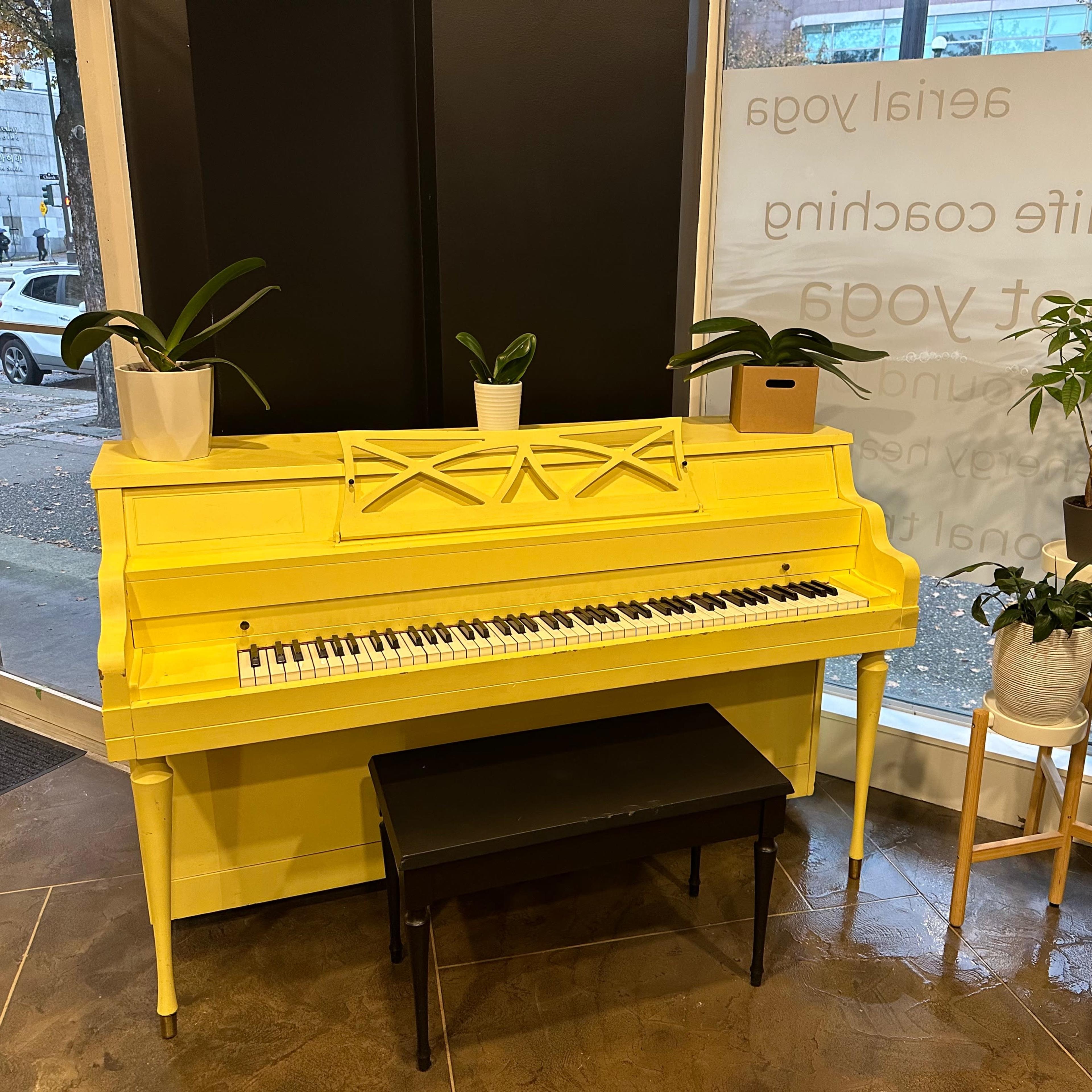 A yellow upright piano positioned next to two potted plants and a black bench in a well-lit interior space.