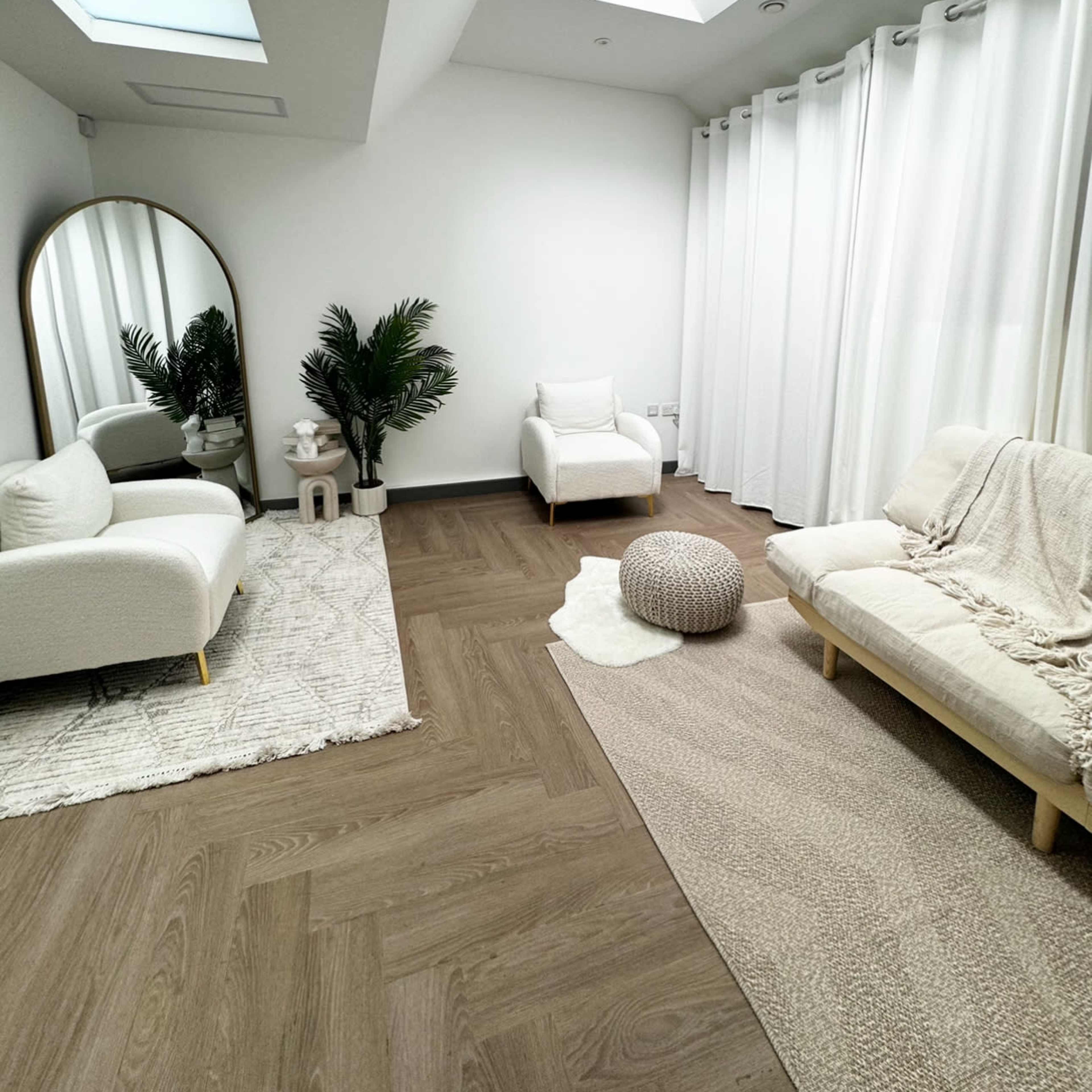 A modern living space with white furniture, large windows, and light wood flooring, featuring a mirror, a potted plant, and various textiles.