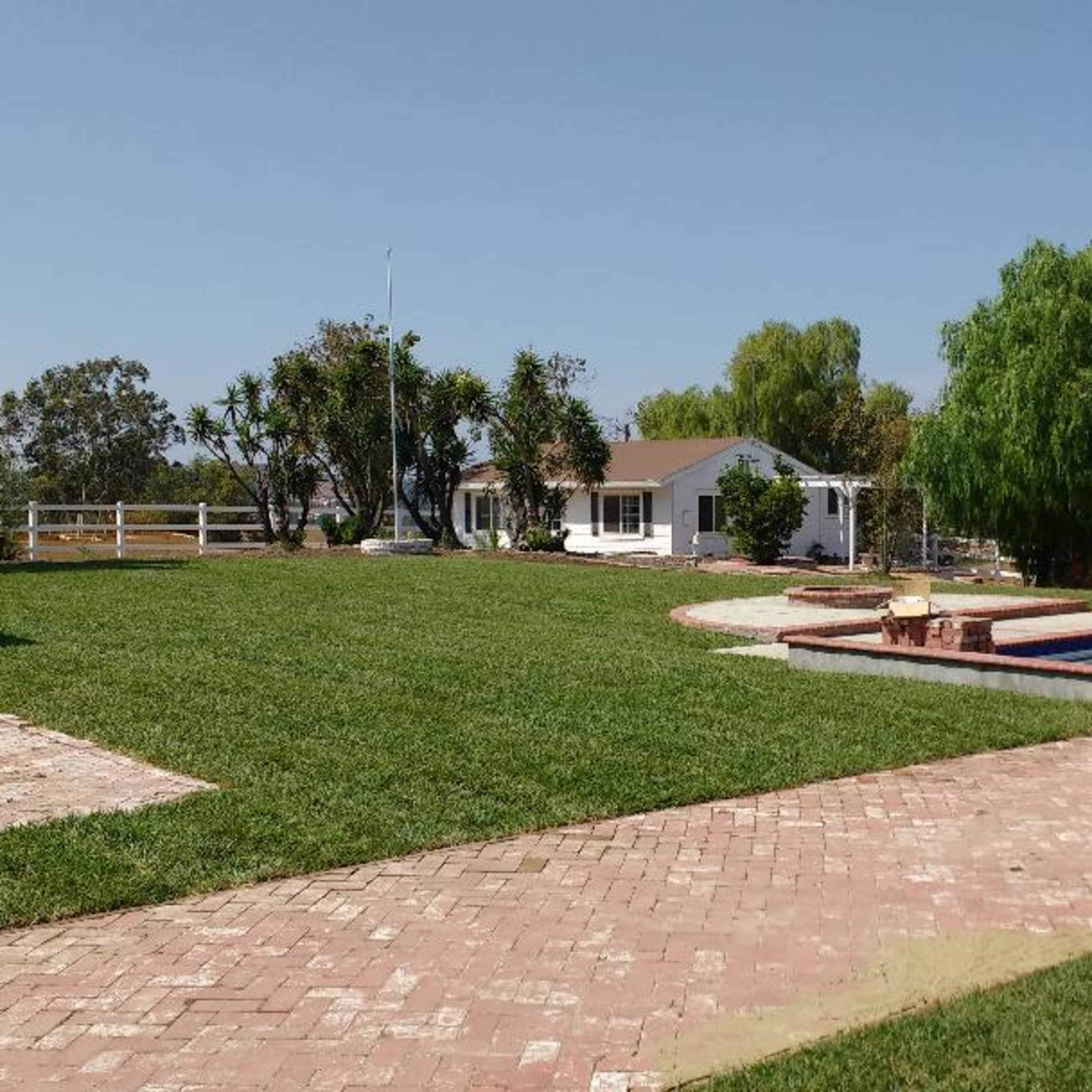 A spacious backyard featuring a swimming pool, a brick walkway, and several trees, with a house visible in the background.
