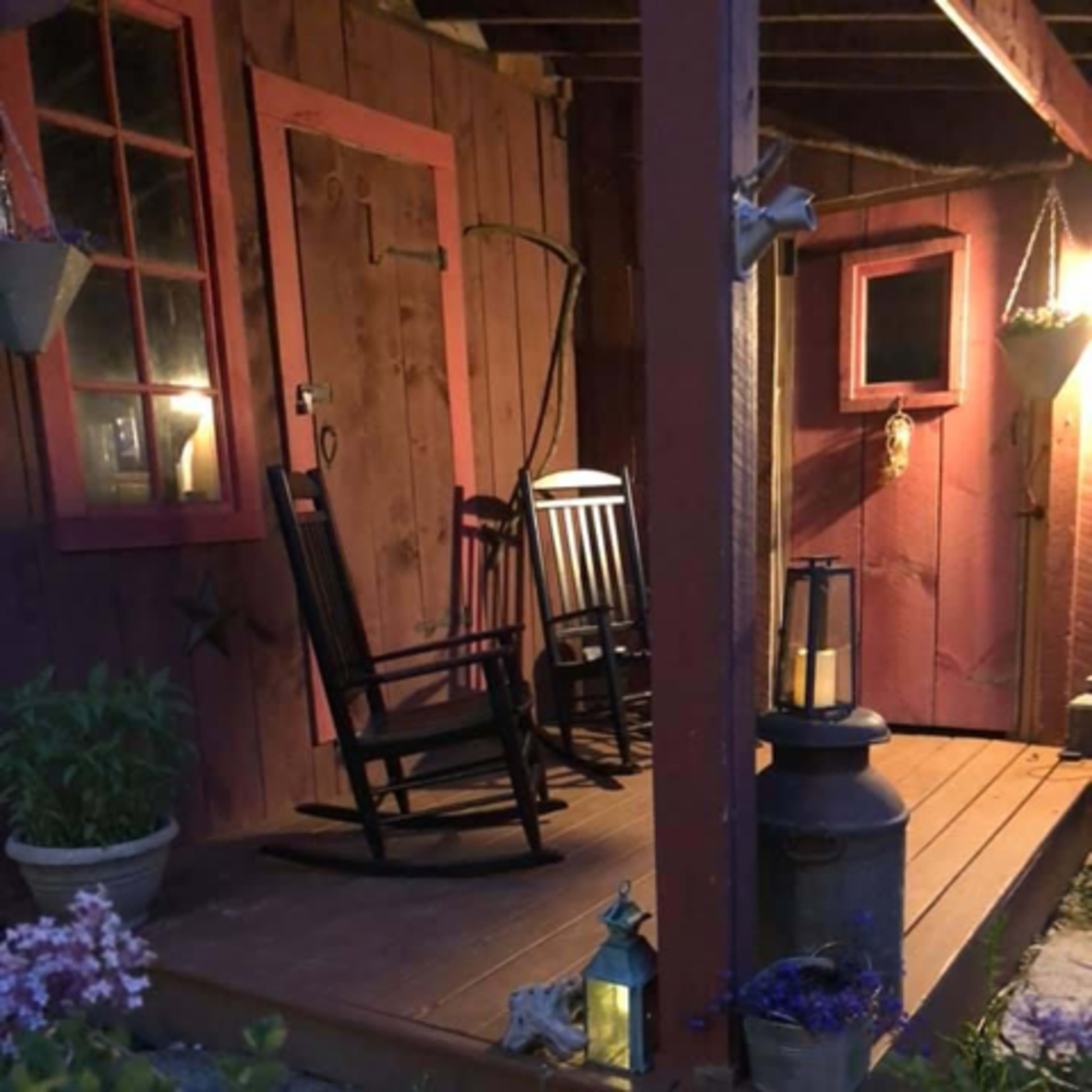 A rustic porch featuring two rocking chairs, potted plants, lanterns, and wooden walls with red trim.