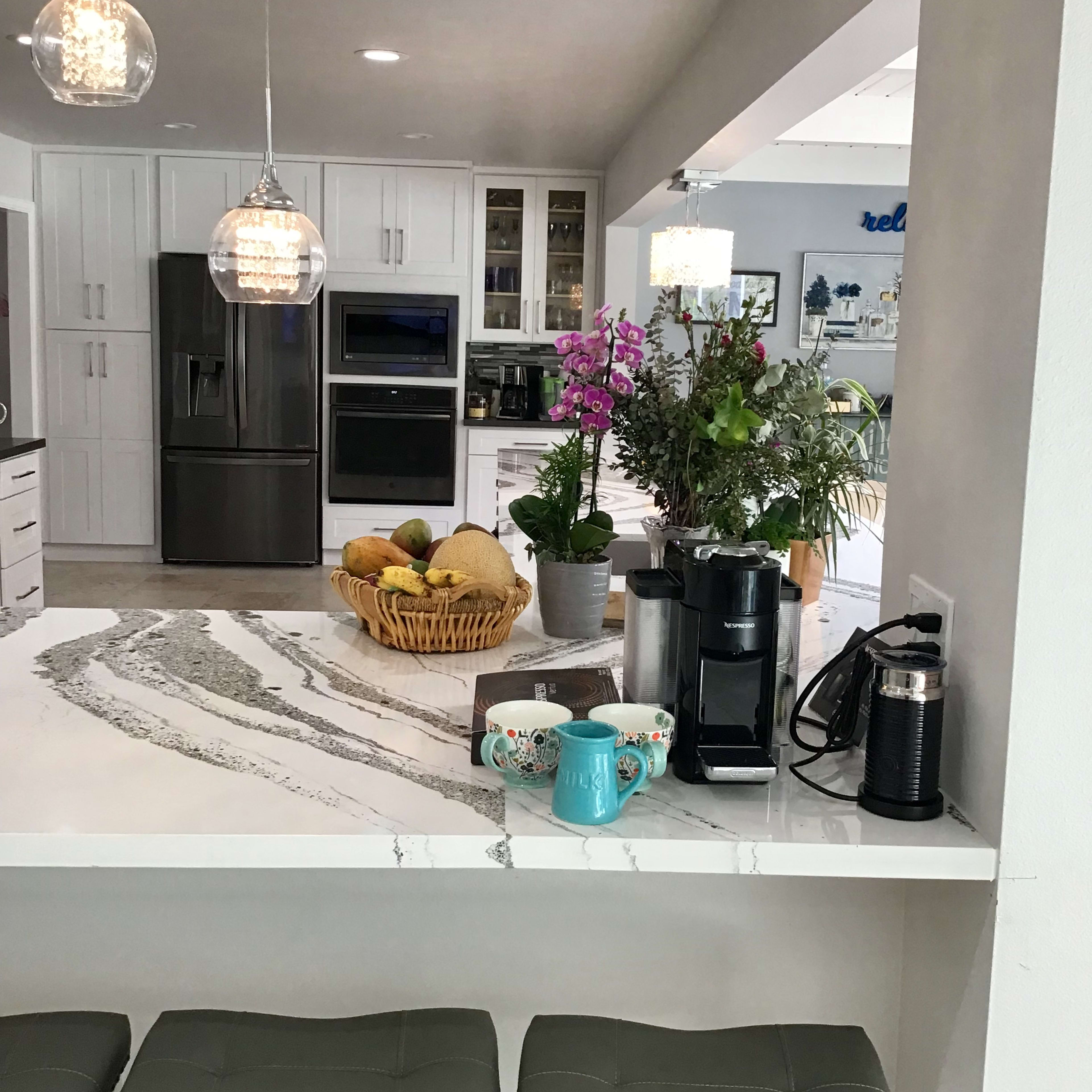 A modern kitchen features a marble countertop with a coffee maker and colorful mugs, surrounded by pendant lighting and various plants.