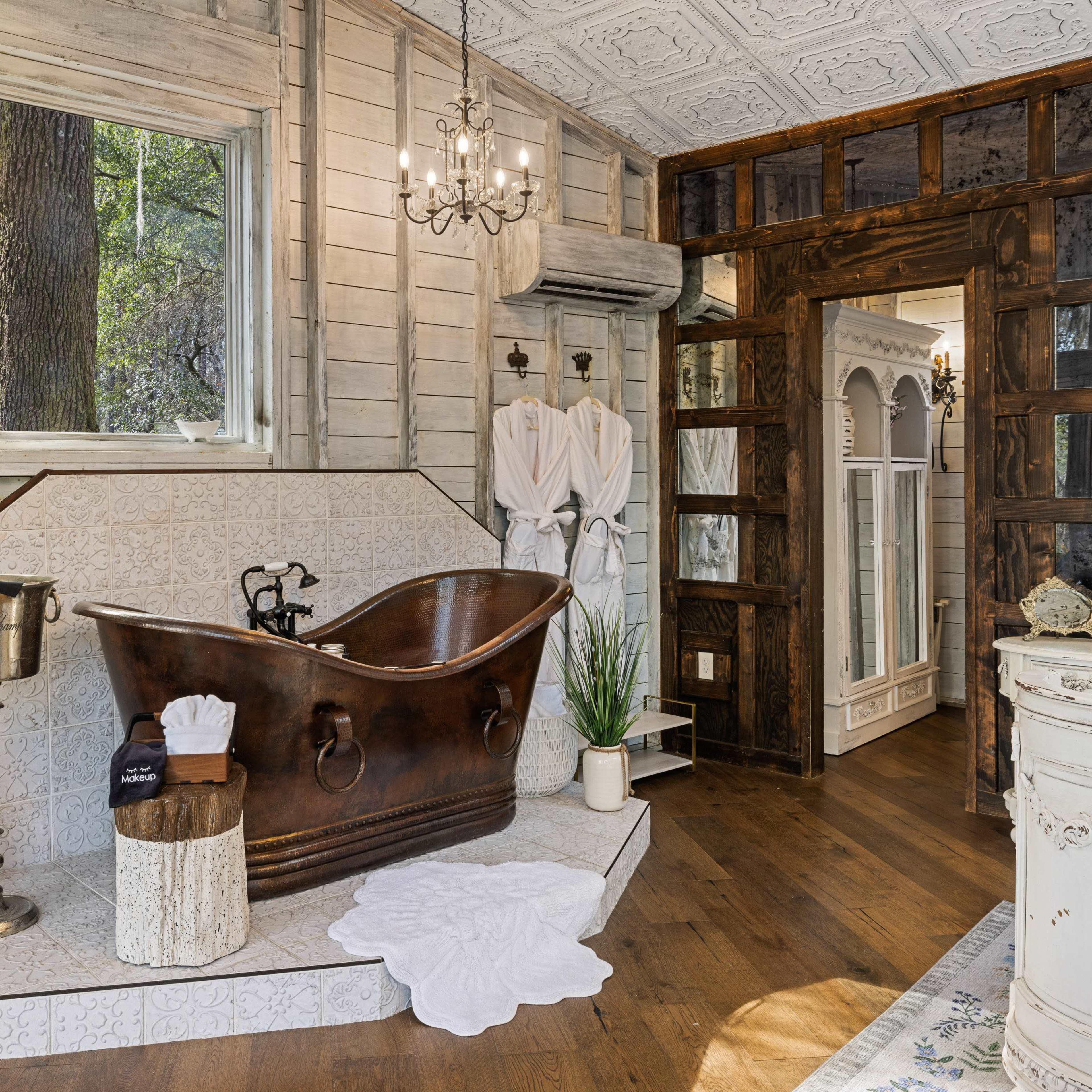 A rustic bathroom featuring a large, freestanding copper bathtub, wooden accents, and a large window with a view of trees outside.