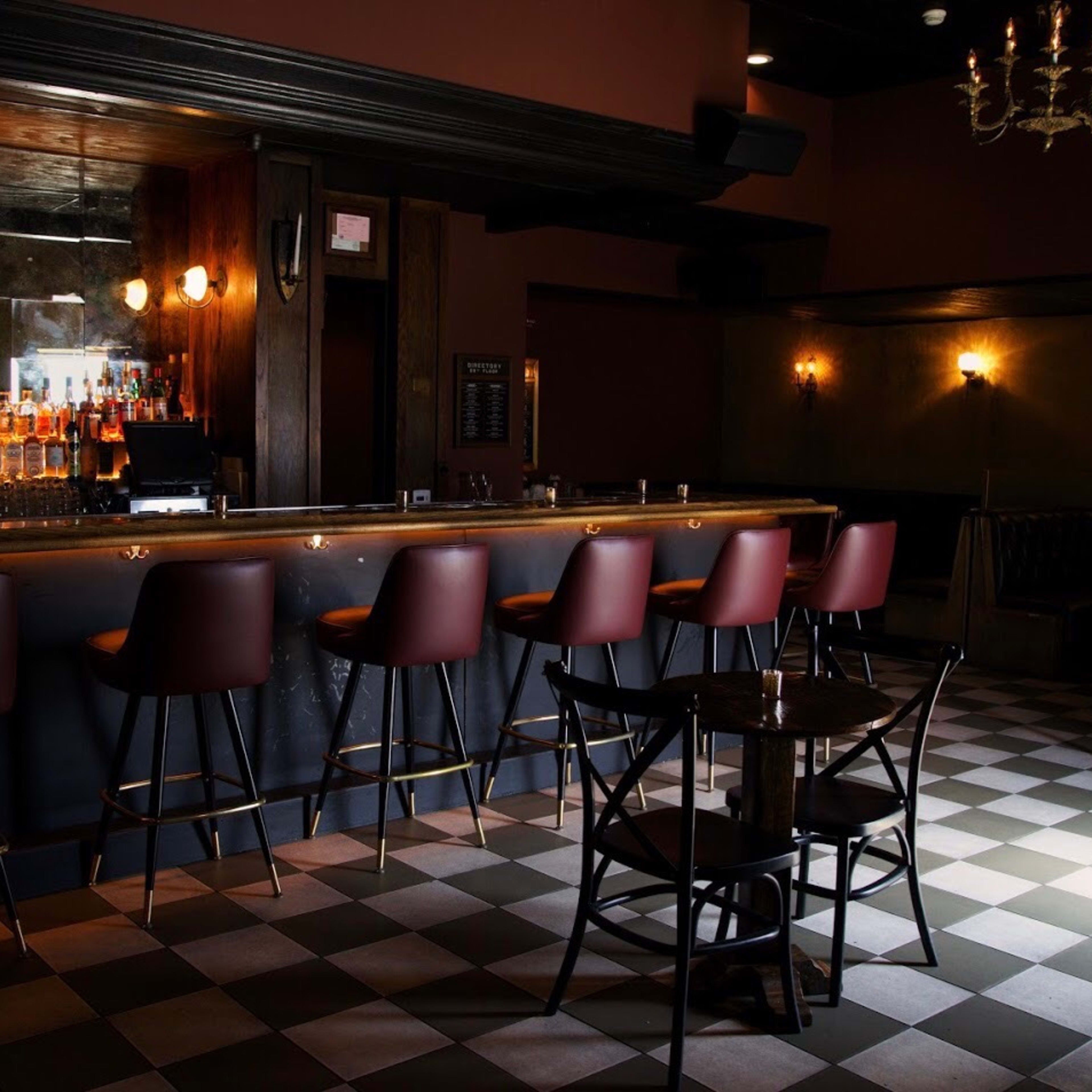 A dimly lit bar area with a checkerboard floor, a long counter lined with high stools, and shelves stocked with liquor bottles in the background.
