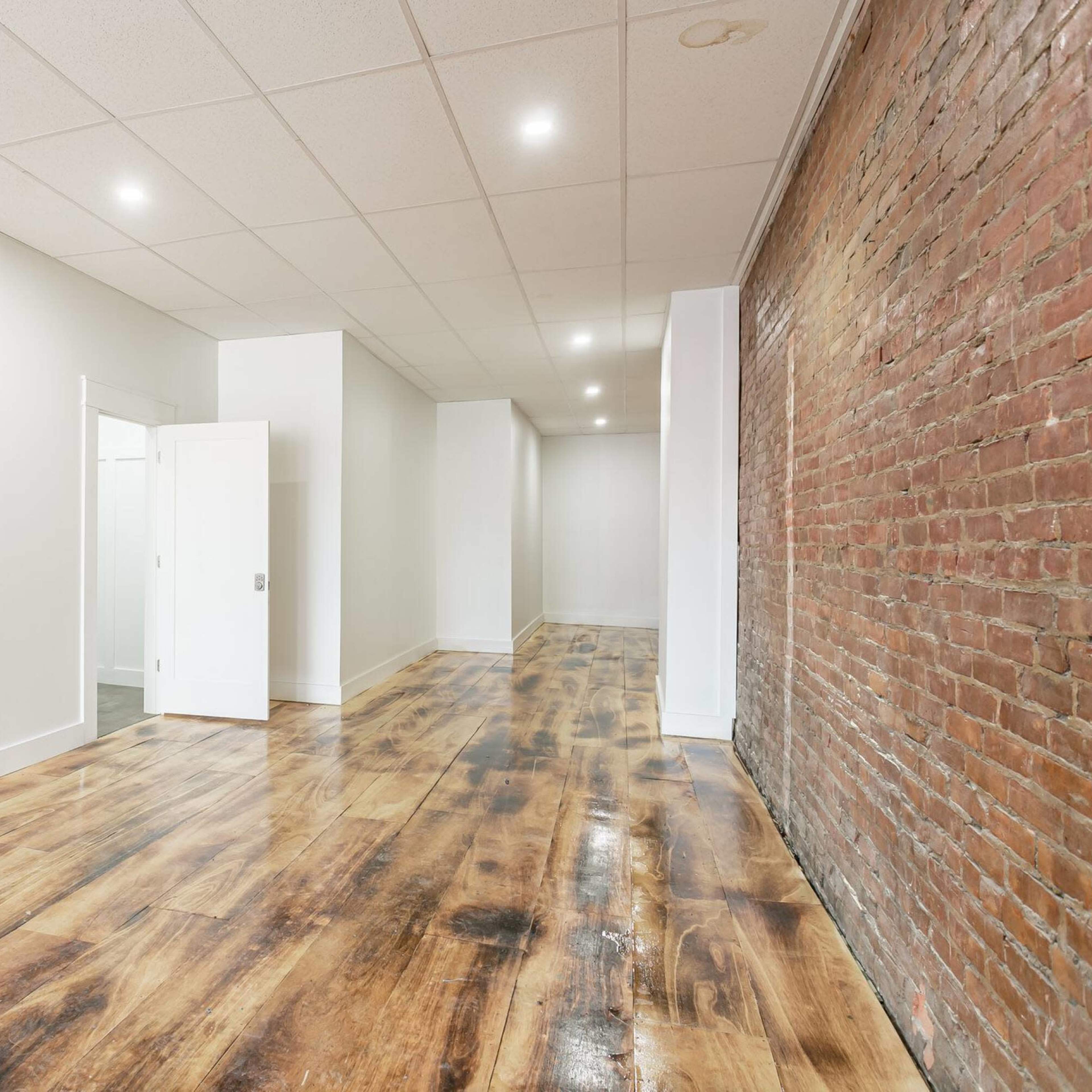 A spacious interior with a polished wooden floor, a brick accent wall, and white walls illuminated by recessed lighting.