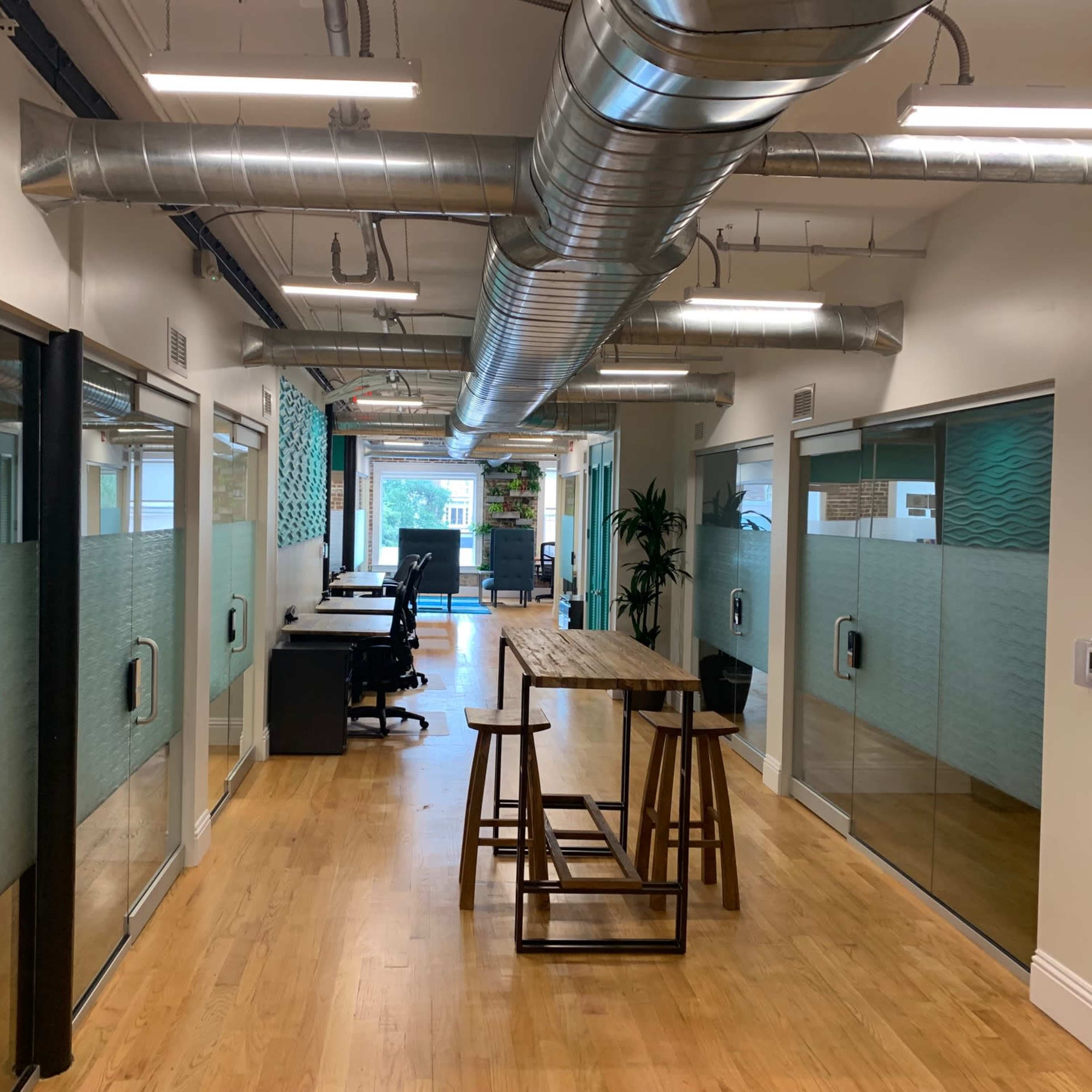 A modern office corridor with glass meeting rooms on either side and a communal table in the center.