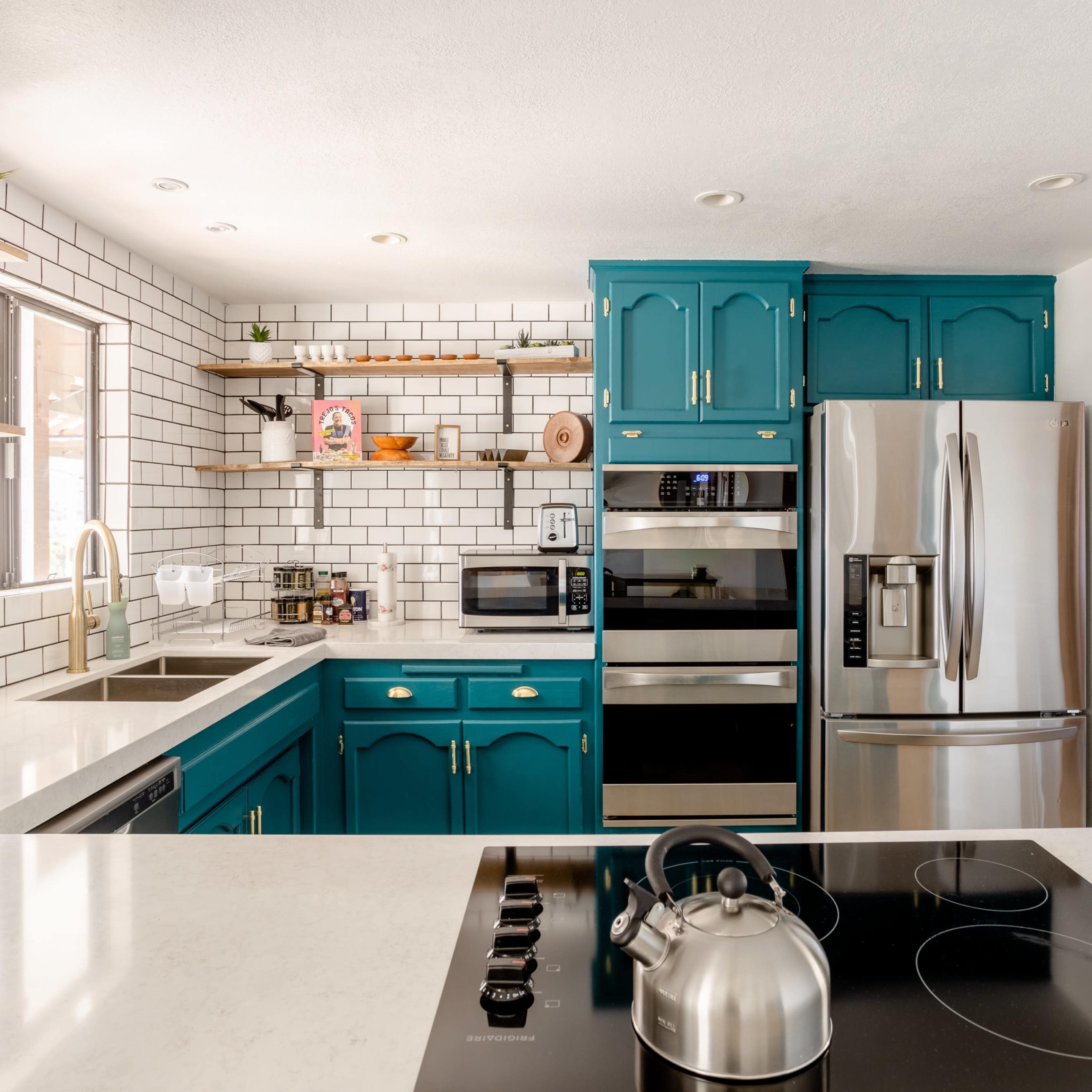 A modern kitchen with teal cabinets, white subway tile backsplashes, stainless steel appliances, and a large island with a stovetop.