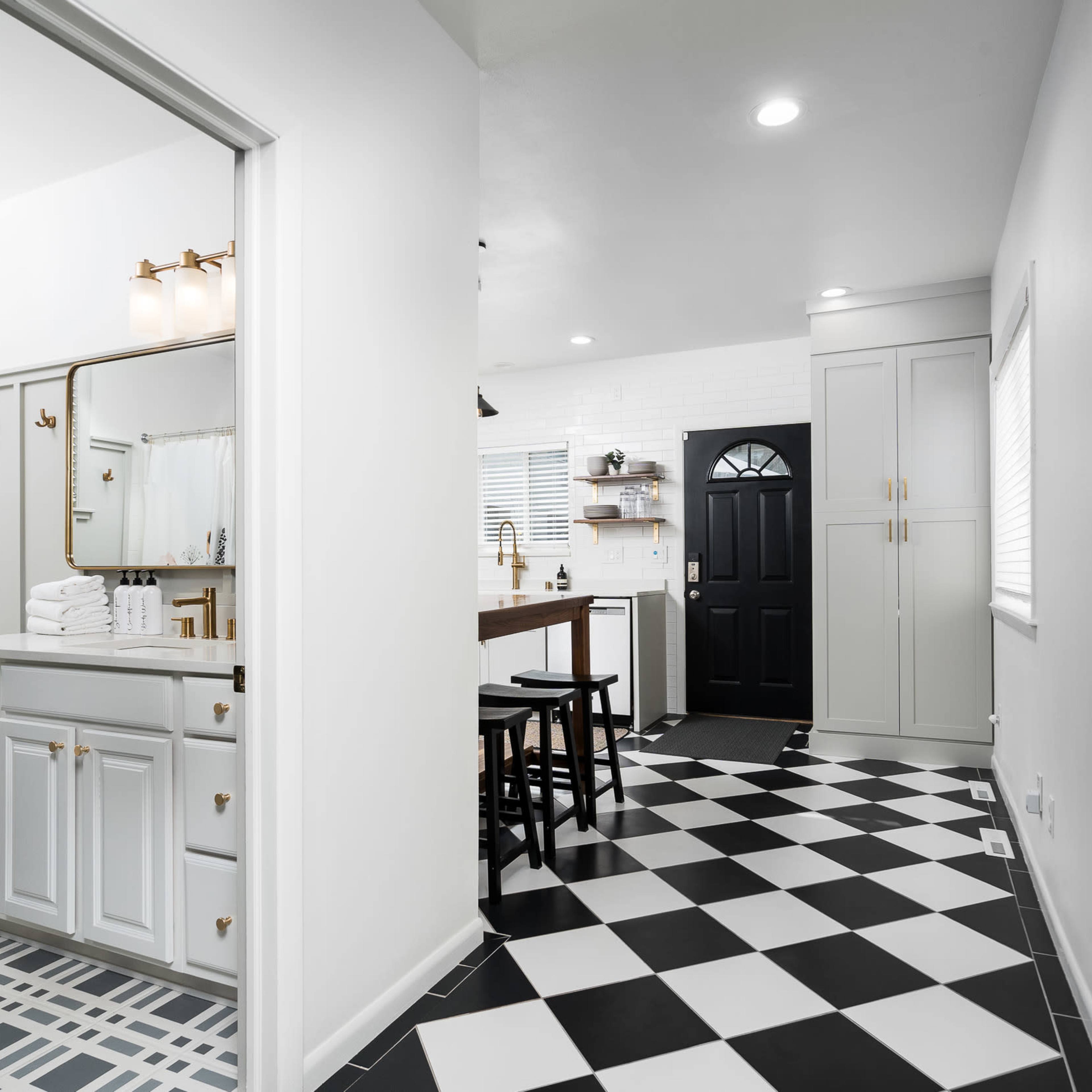 A modern bathroom with a white vanity and black and white tiled flooring, leading into a hallway and kitchen area with a black front door.