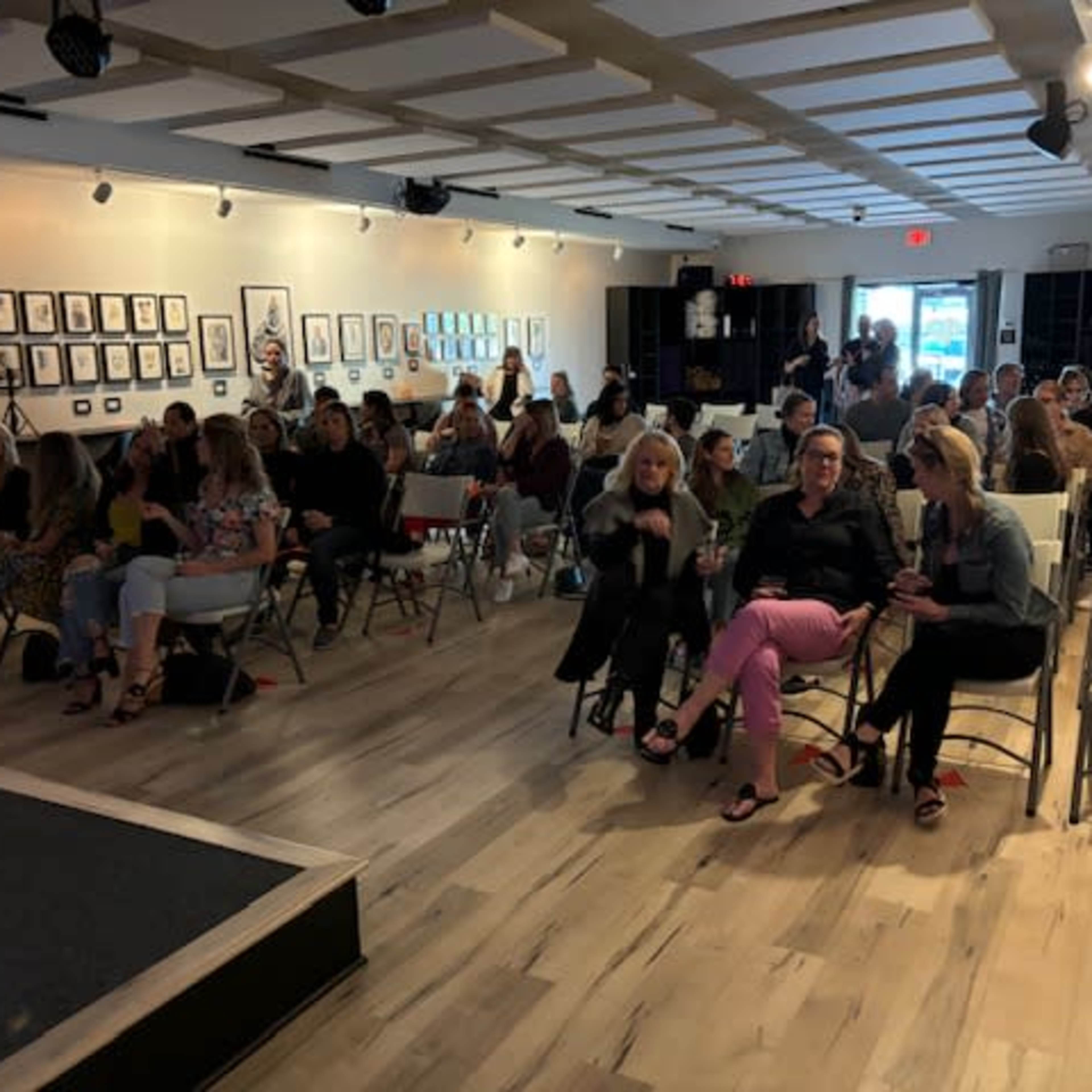 A seated audience fills a well-lit room adorned with framed artwork, waiting for an event to begin.