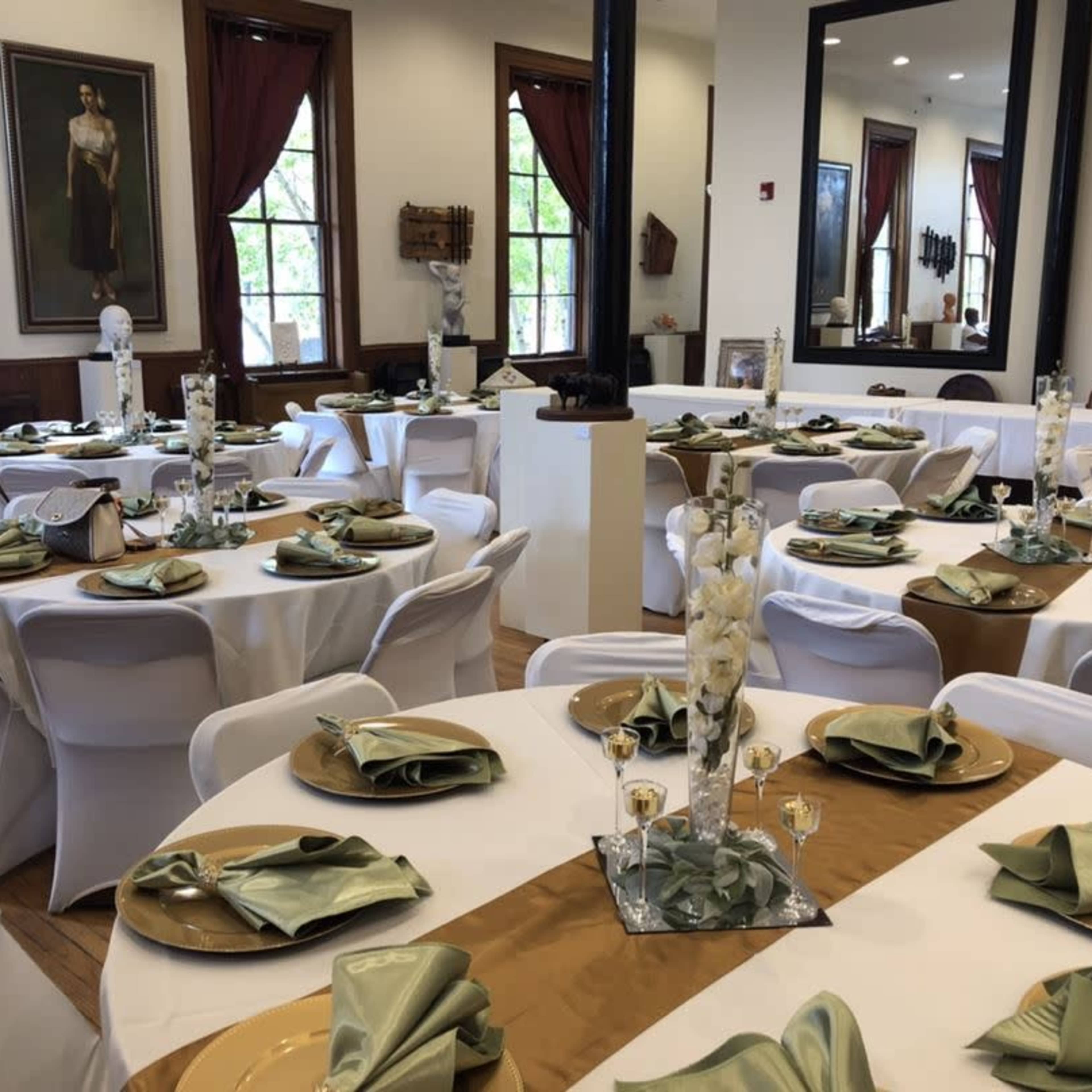 A banquet hall set with round tables that are covered with white tablecloths and adorned with green napkins and gold plates.