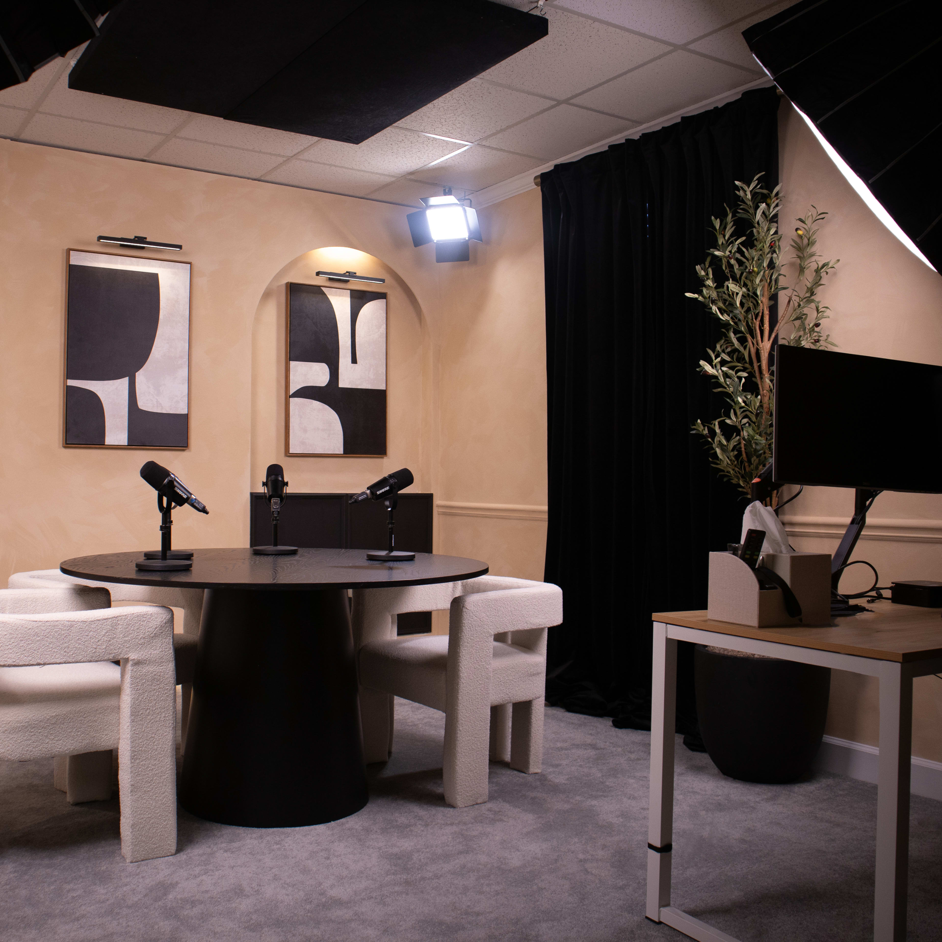 A modern recording studio featuring a round black table with two microphones, three white upholstered chairs, and wall art, set against a tan-colored backdrop.