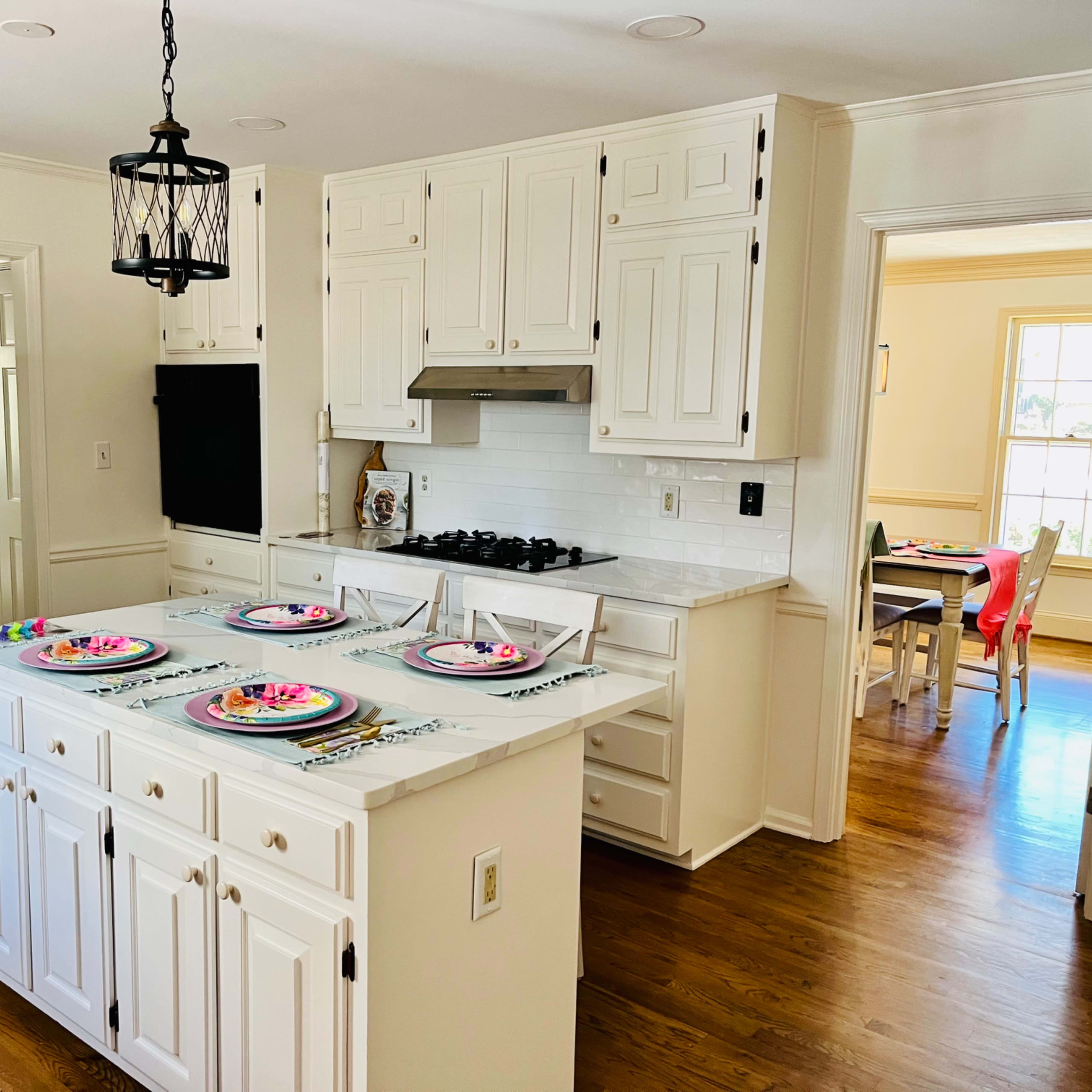 A well-lit kitchen features white cabinetry, a central island with colorful plates set for four, and an adjacent dining area visible through an open doorway.