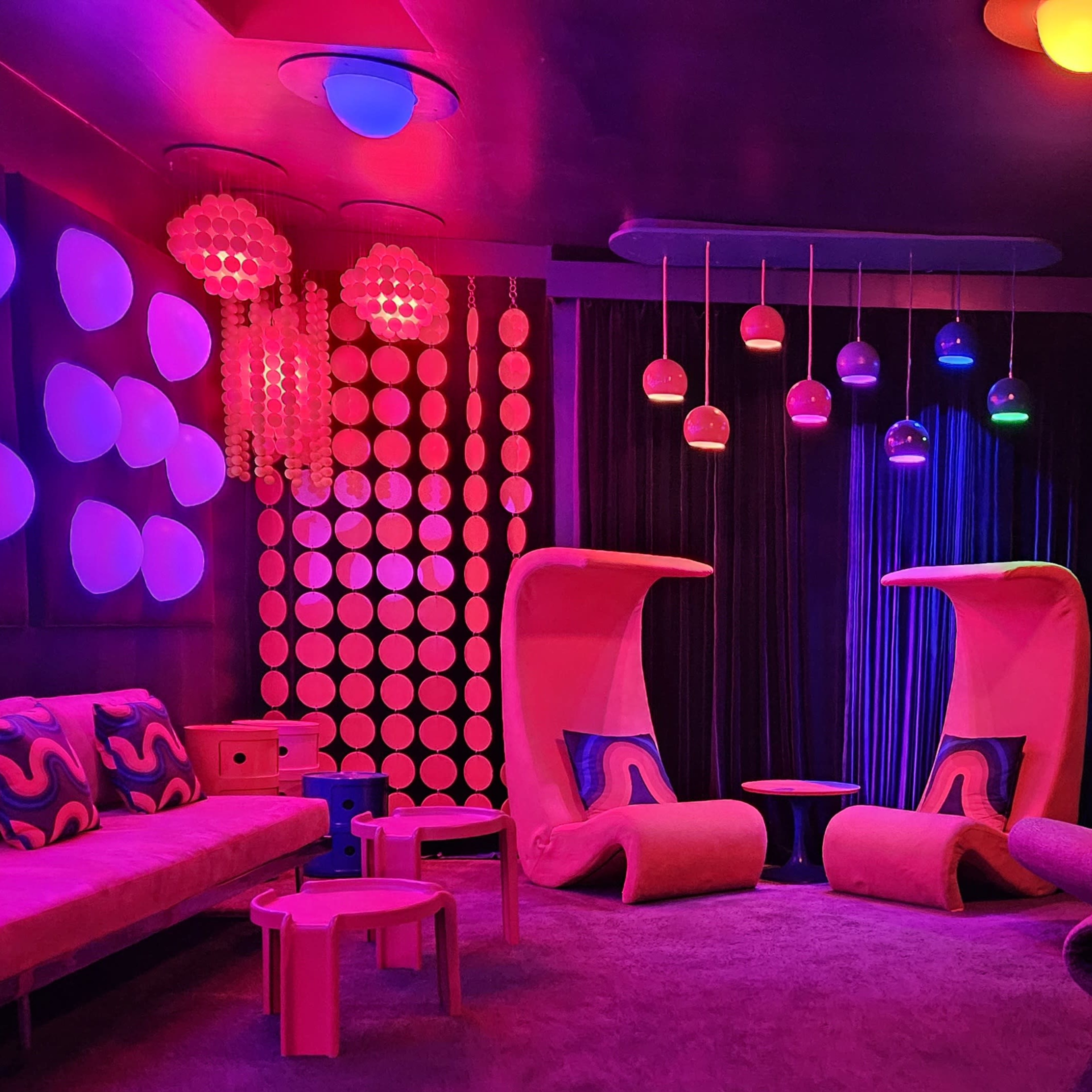 A vibrant, retro-themed lounge with colorful furniture, including large chairs and wall decorations featuring polka dots, illuminated by various pendant lights.