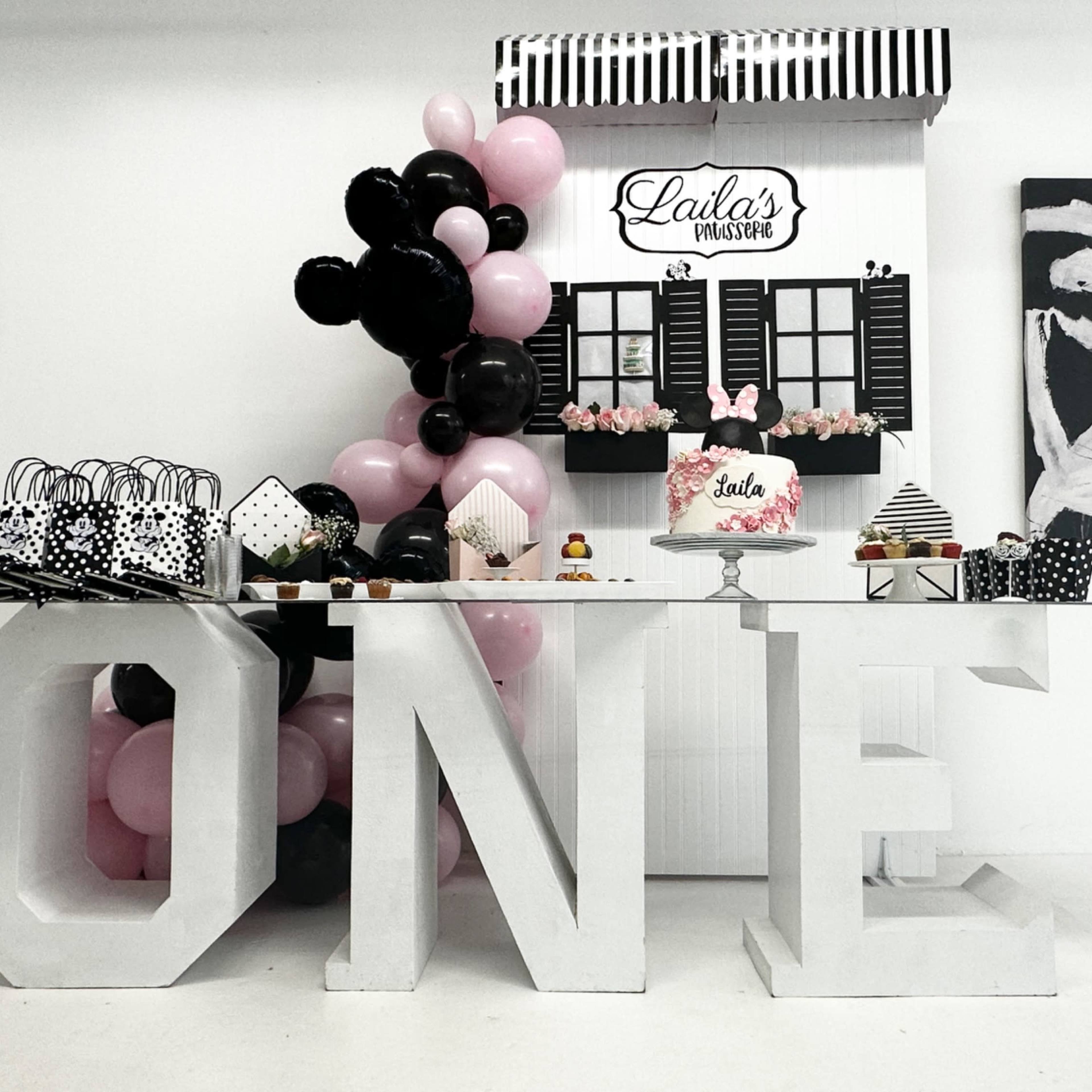 A decorated table set up for a one-year-old's birthday party, displaying various treats and a large "ONE" sign, along with black, pink, and white decorations.