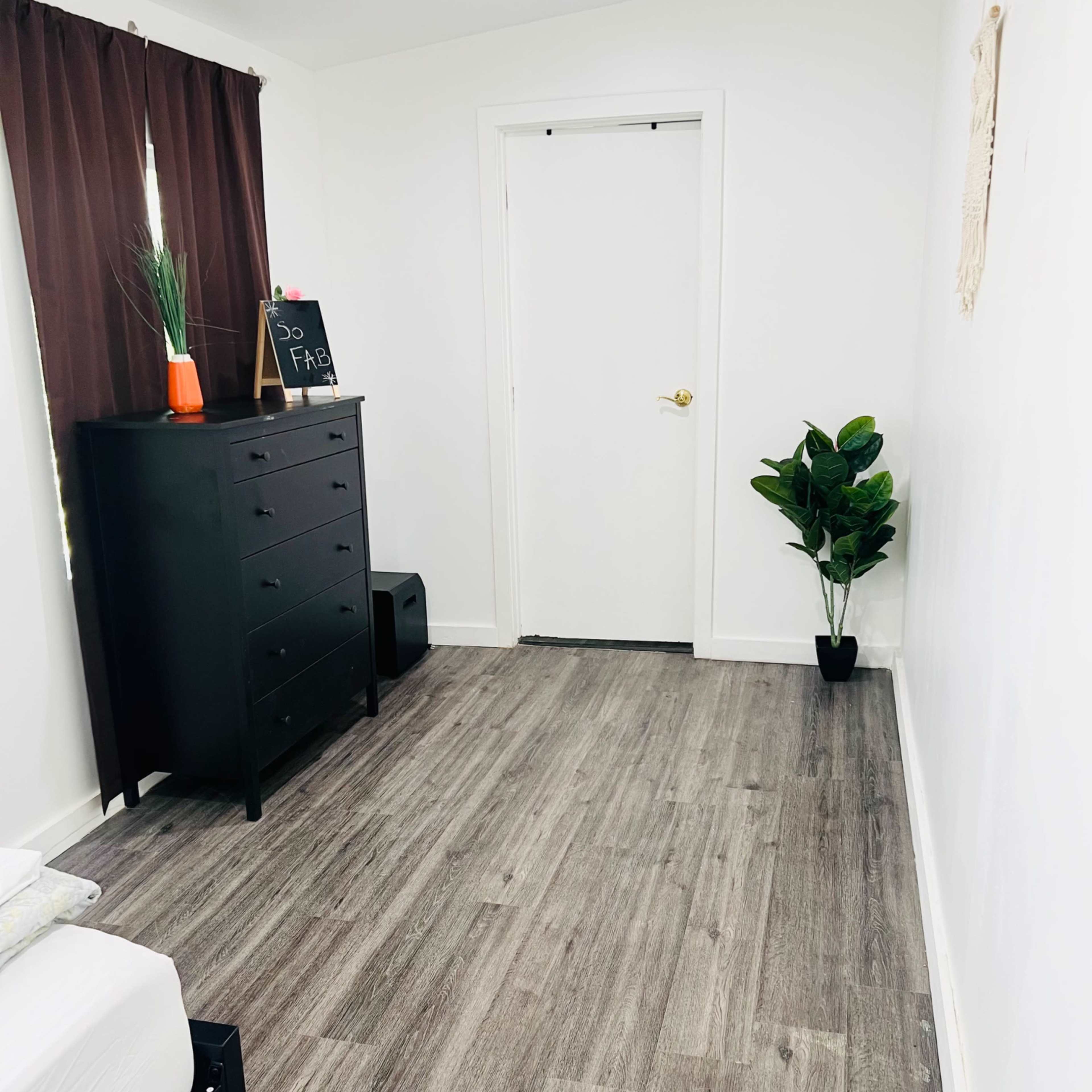 A minimalistic room featuring a dresser, a tall potted plant, and a closed white door against a wall with light-colored flooring.