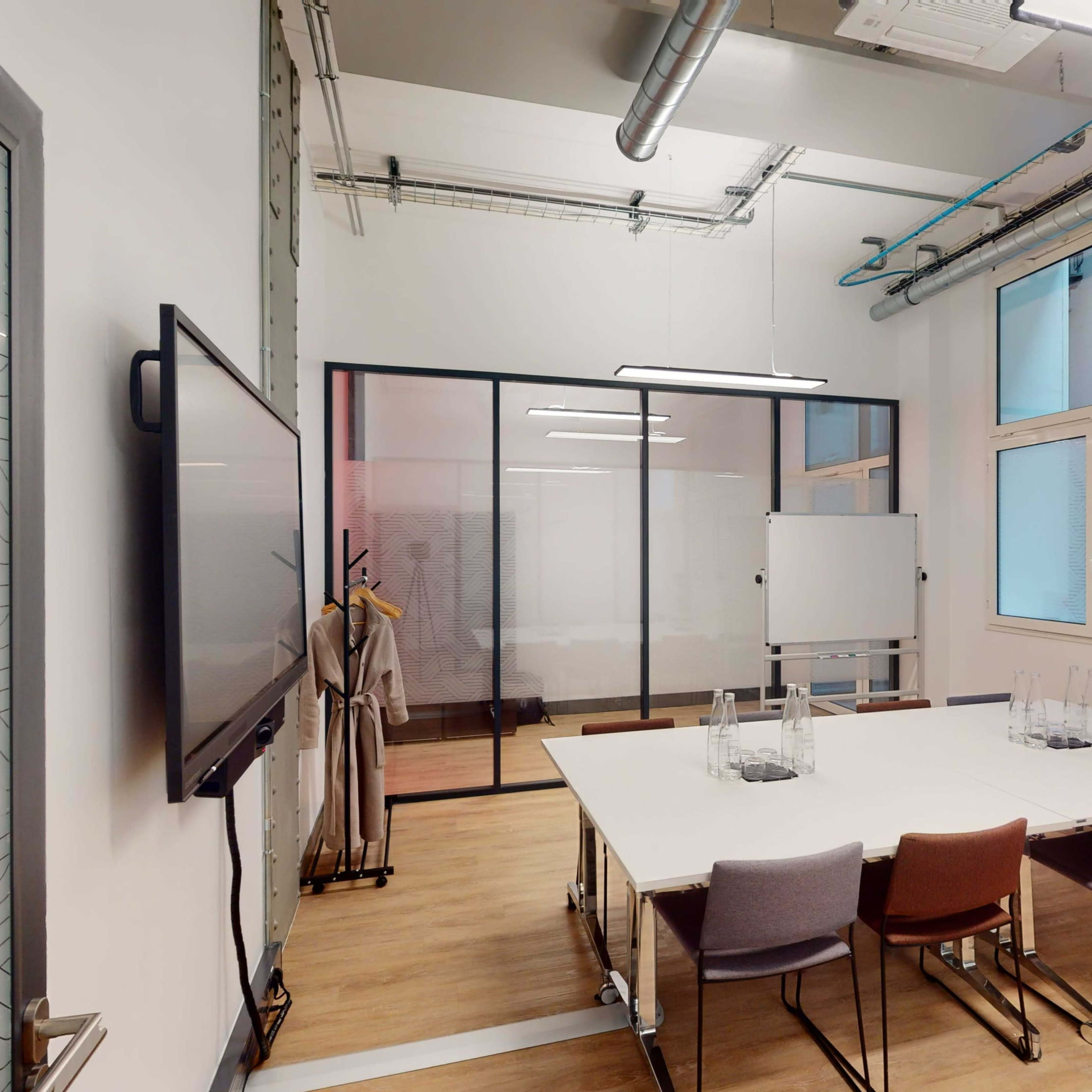 A modern conference room with a large table, six chairs, a screen, and a glass partition separating it from another workspace.