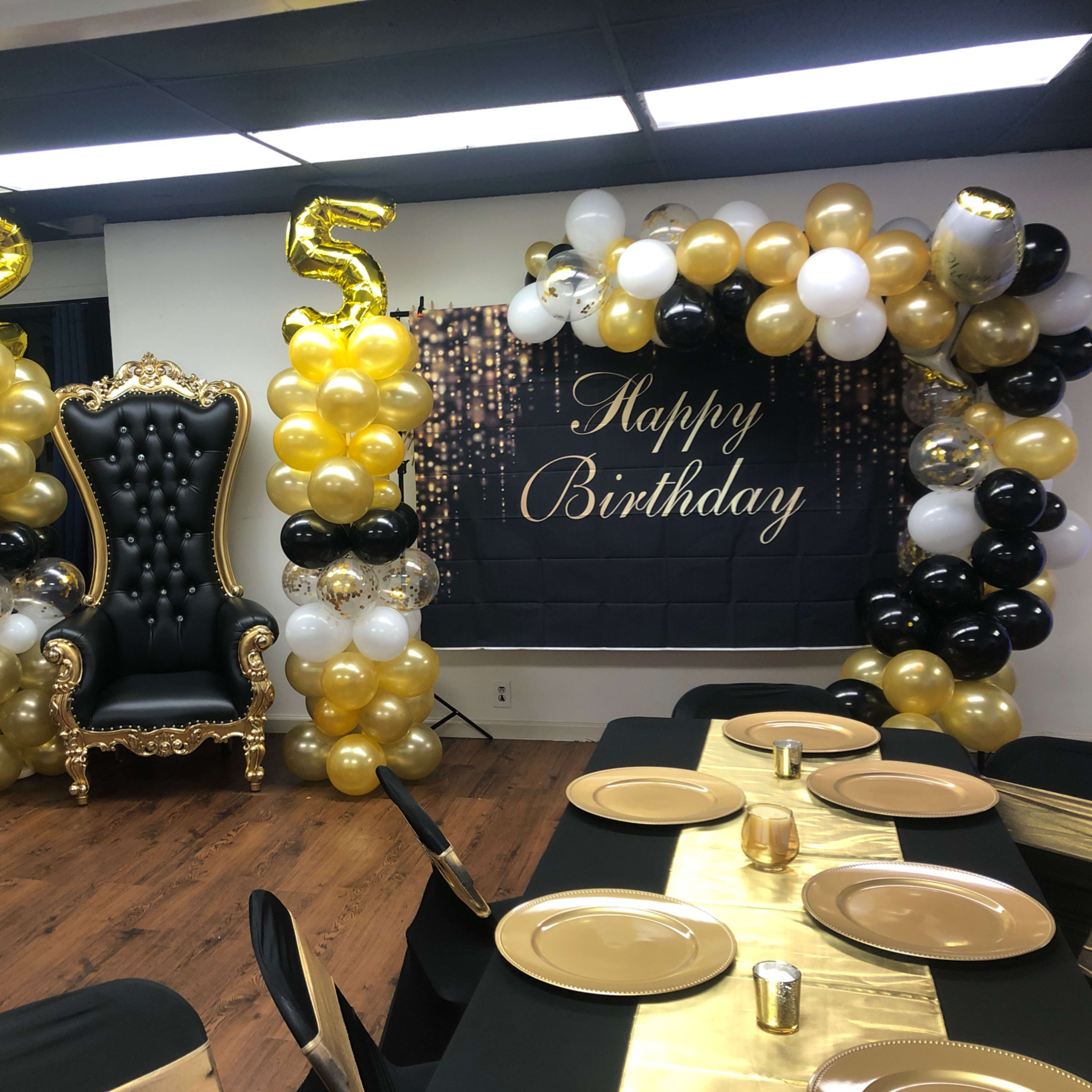A birthday celebration setup featuring a black and gold color scheme, with a large throne chair, an arch of balloons, and a "Happy Birthday" banner.