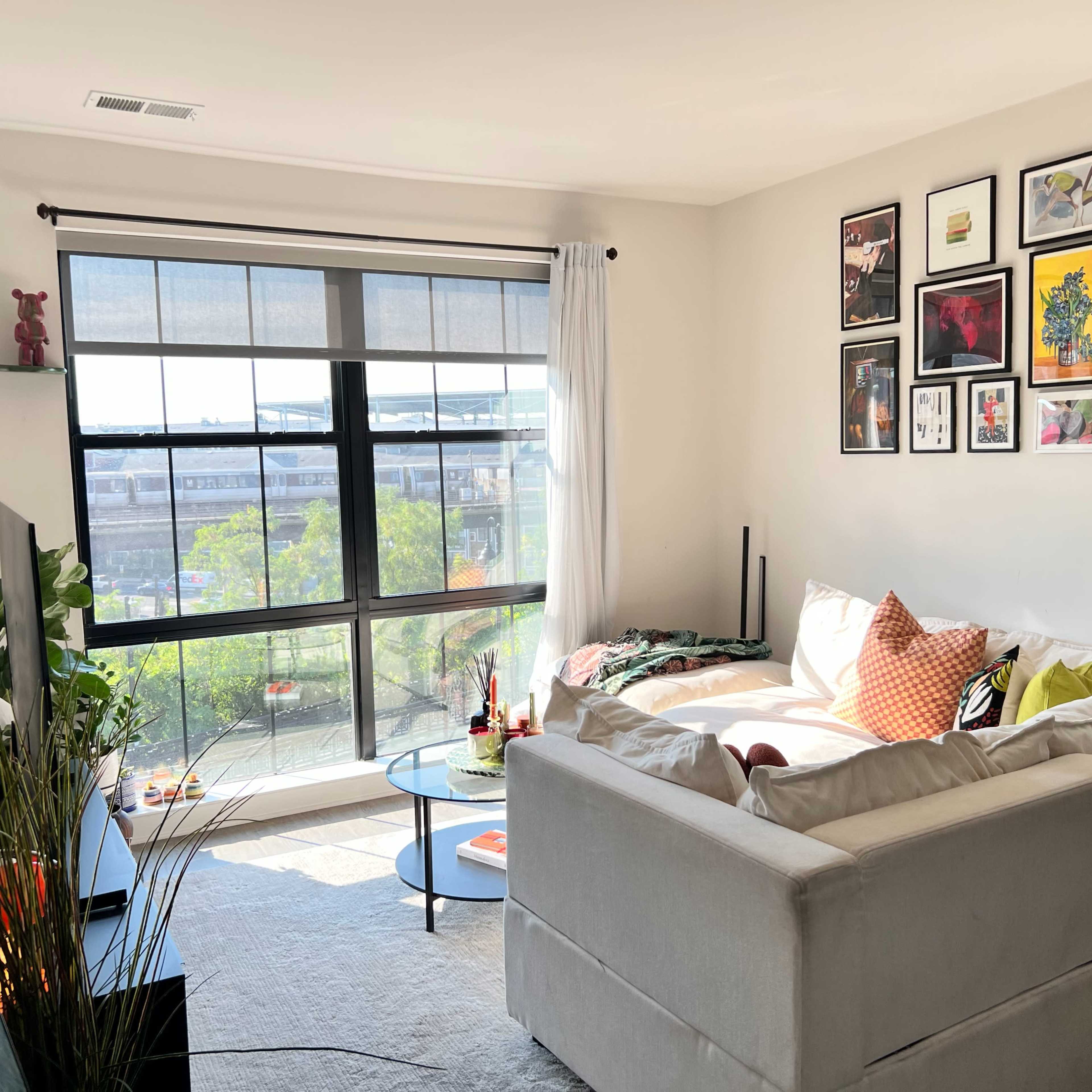 A bright living room features a white sectional sofa, a black coffee table, and a large window with light filtering in, adorned with framed artwork on the walls.