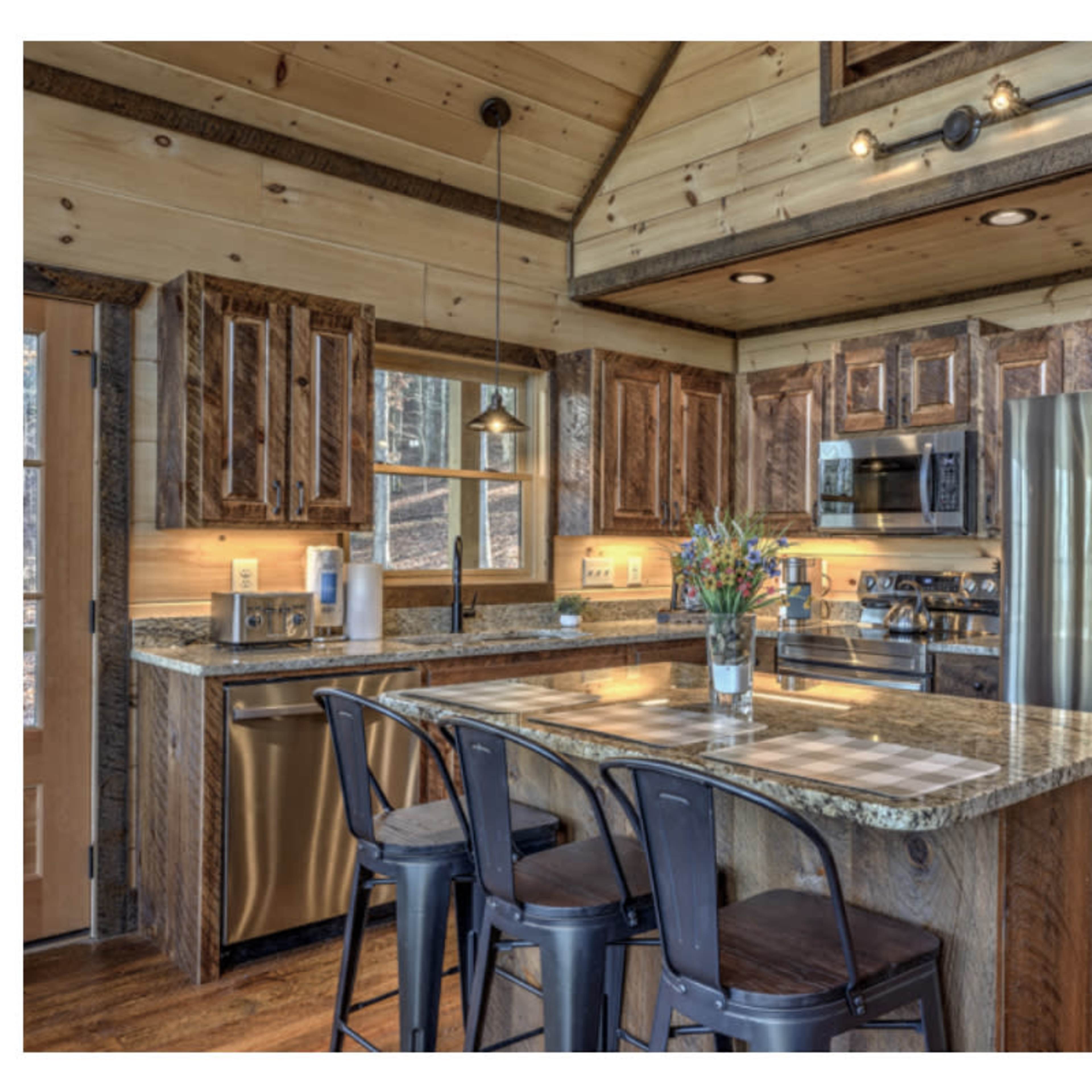 A rustic kitchen featuring wooden cabinets, a granite countertop island with seating, stainless steel appliances, and large windows allowing natural light.