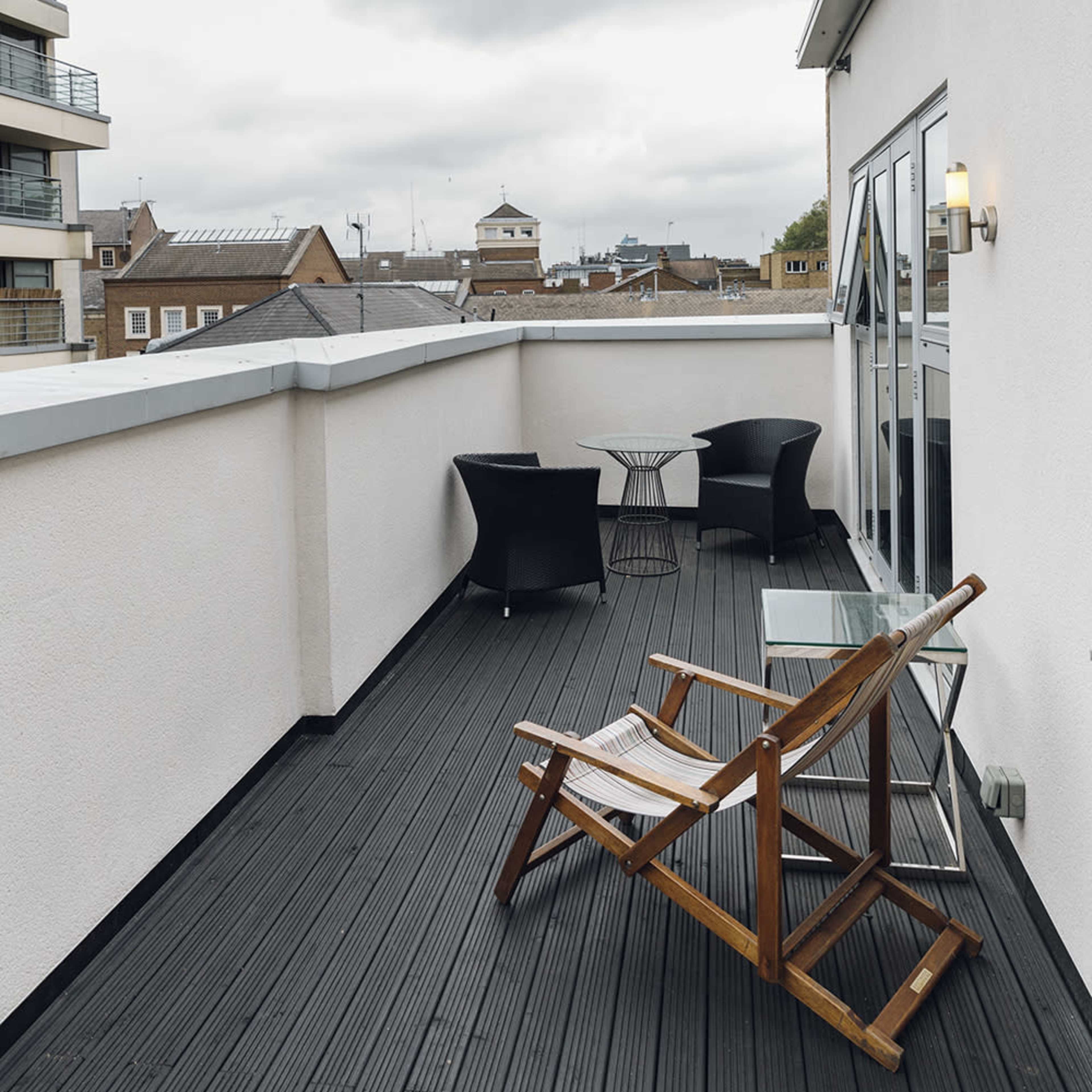 A balcony with a wooden lounge chair, a glass table, and two black chairs, overlooking a cityscape under a cloudy sky.