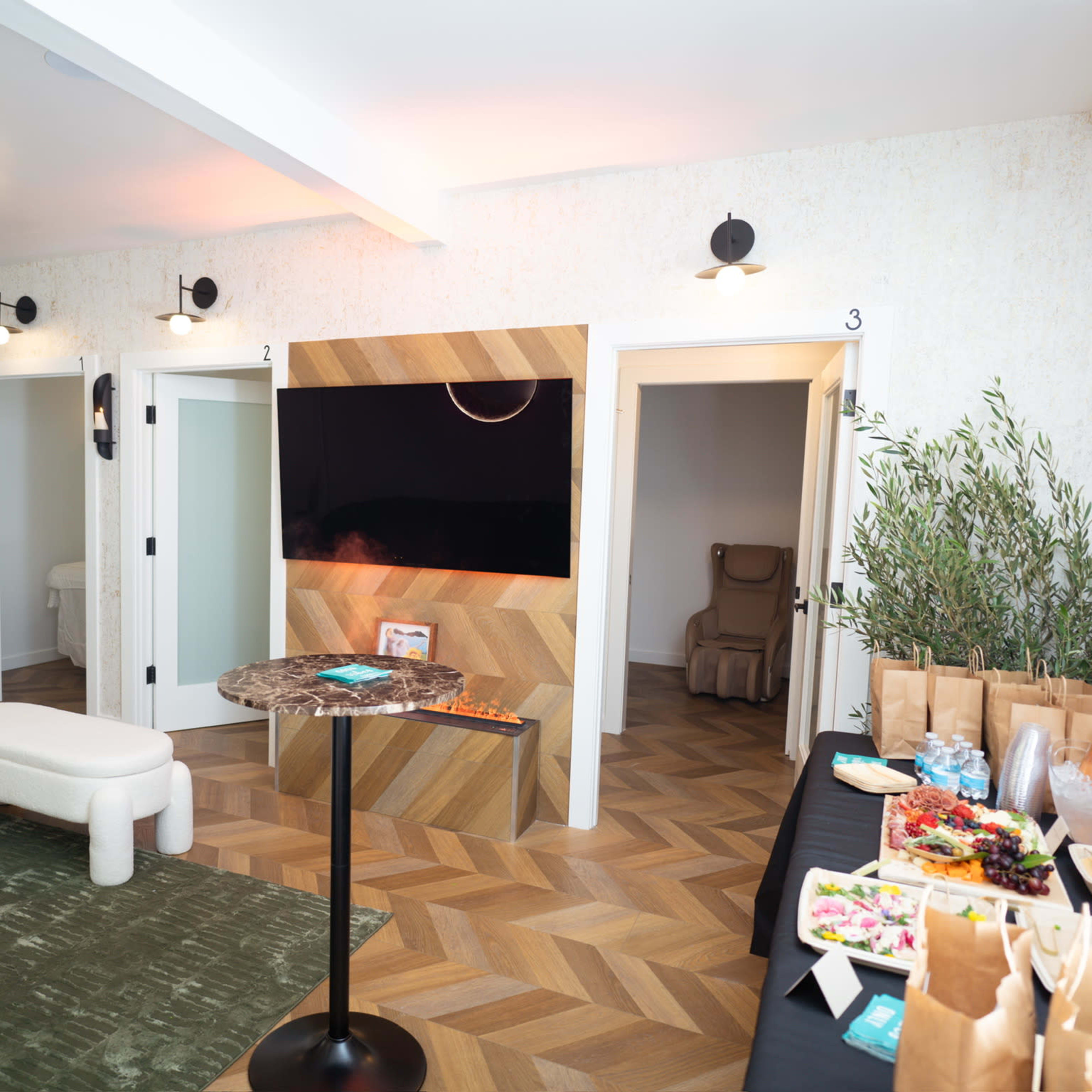 A modern lounge area with a seating arrangement, a TV on a wooden feature wall, three doors leading to other rooms, and a table with food and drinks.