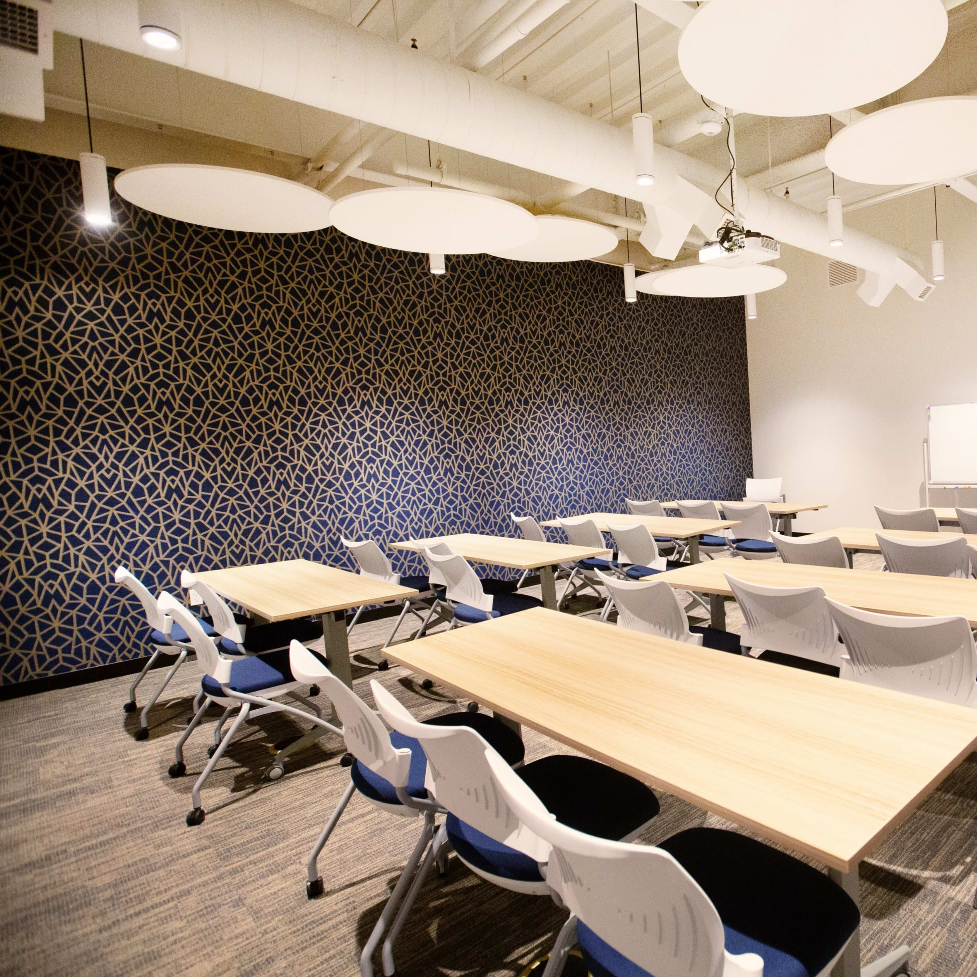 A modern conference room with several tables and chairs arranged for a meeting, featuring a patterned wall and overhead circular light fixtures.