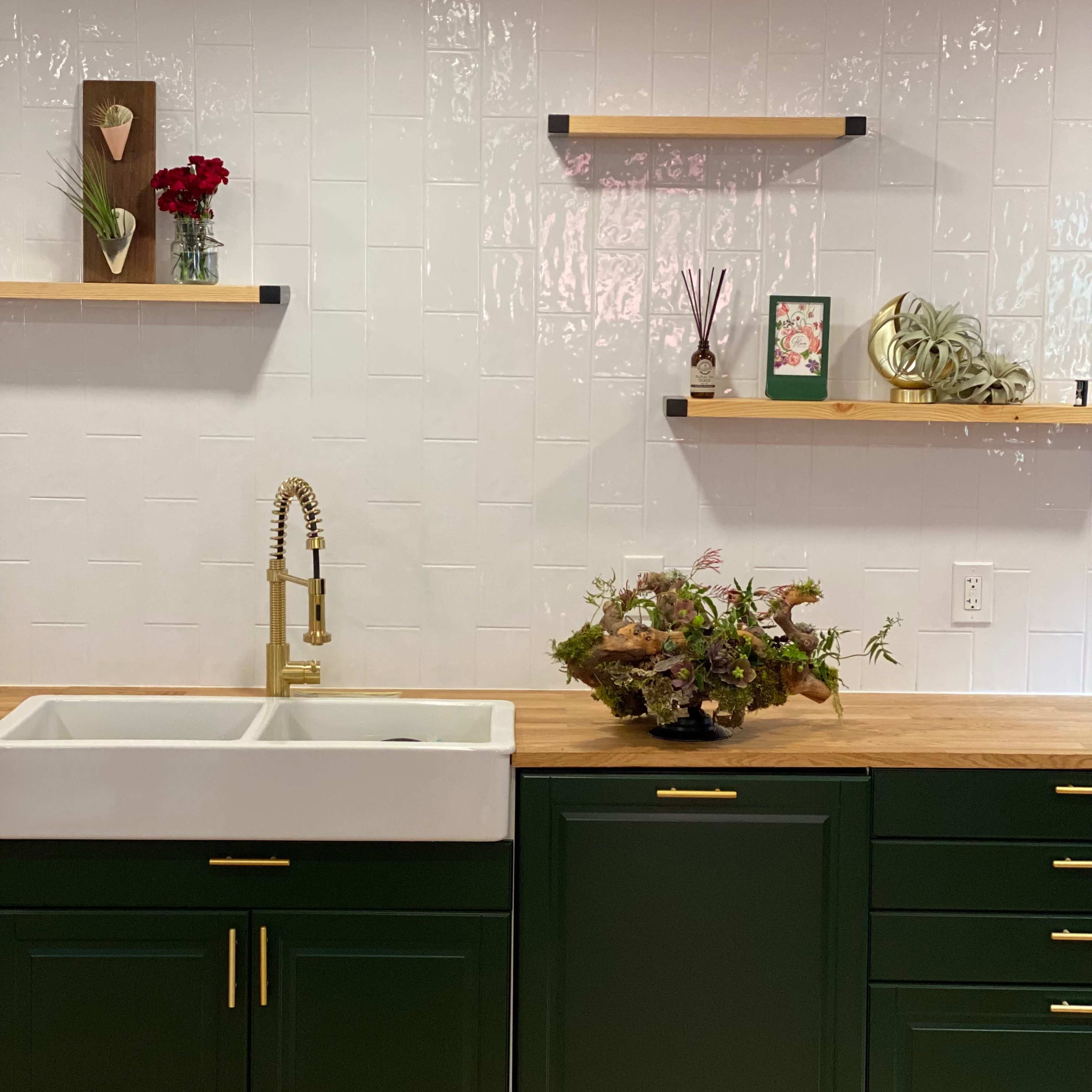 A kitchen with a green base cabinet and a farmhouse sink, accented by wooden shelves displaying decorative items and plants on a white tiled wall.