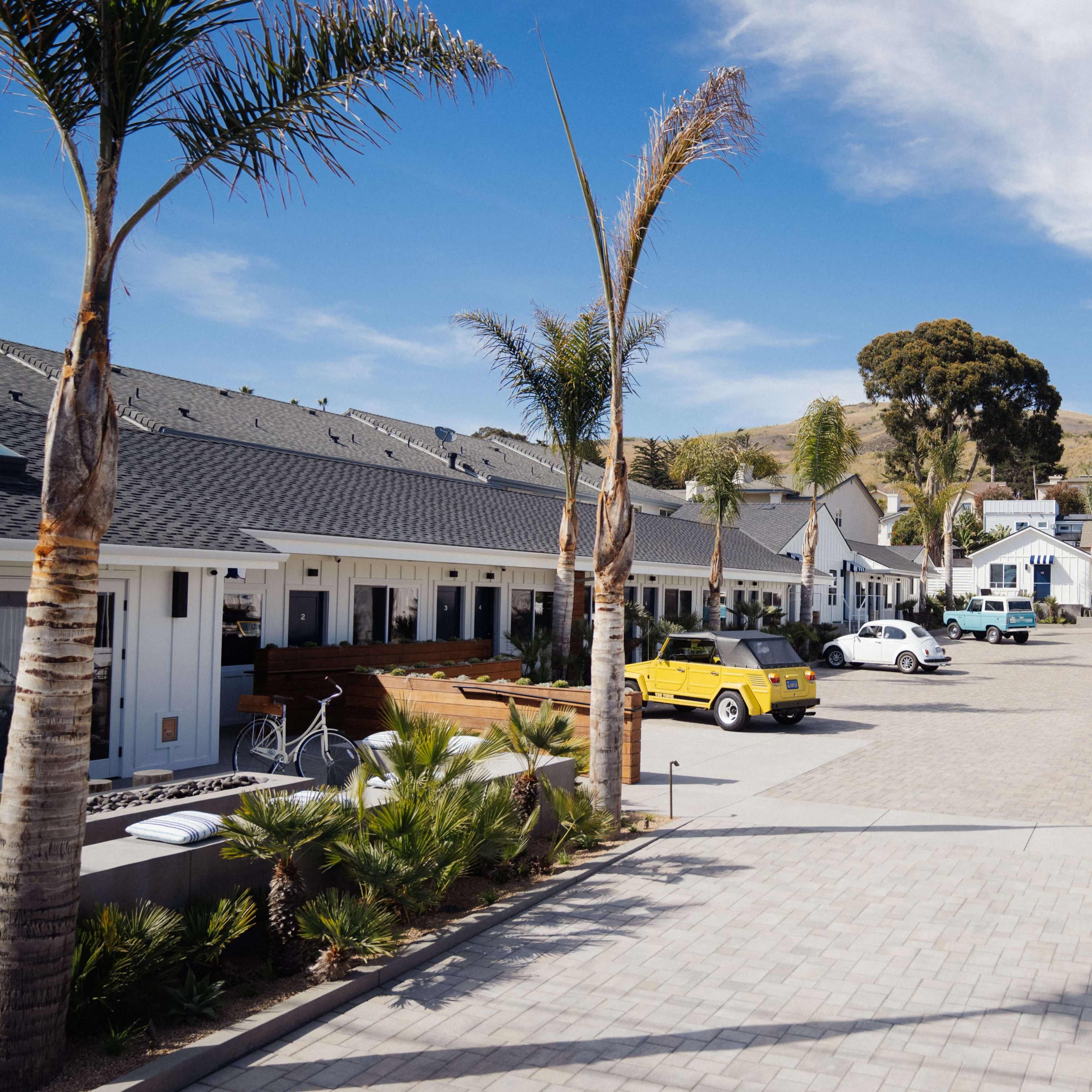 A renovated courtyard with several white cottages lined on either side, palm trees, and a range of parked cars.