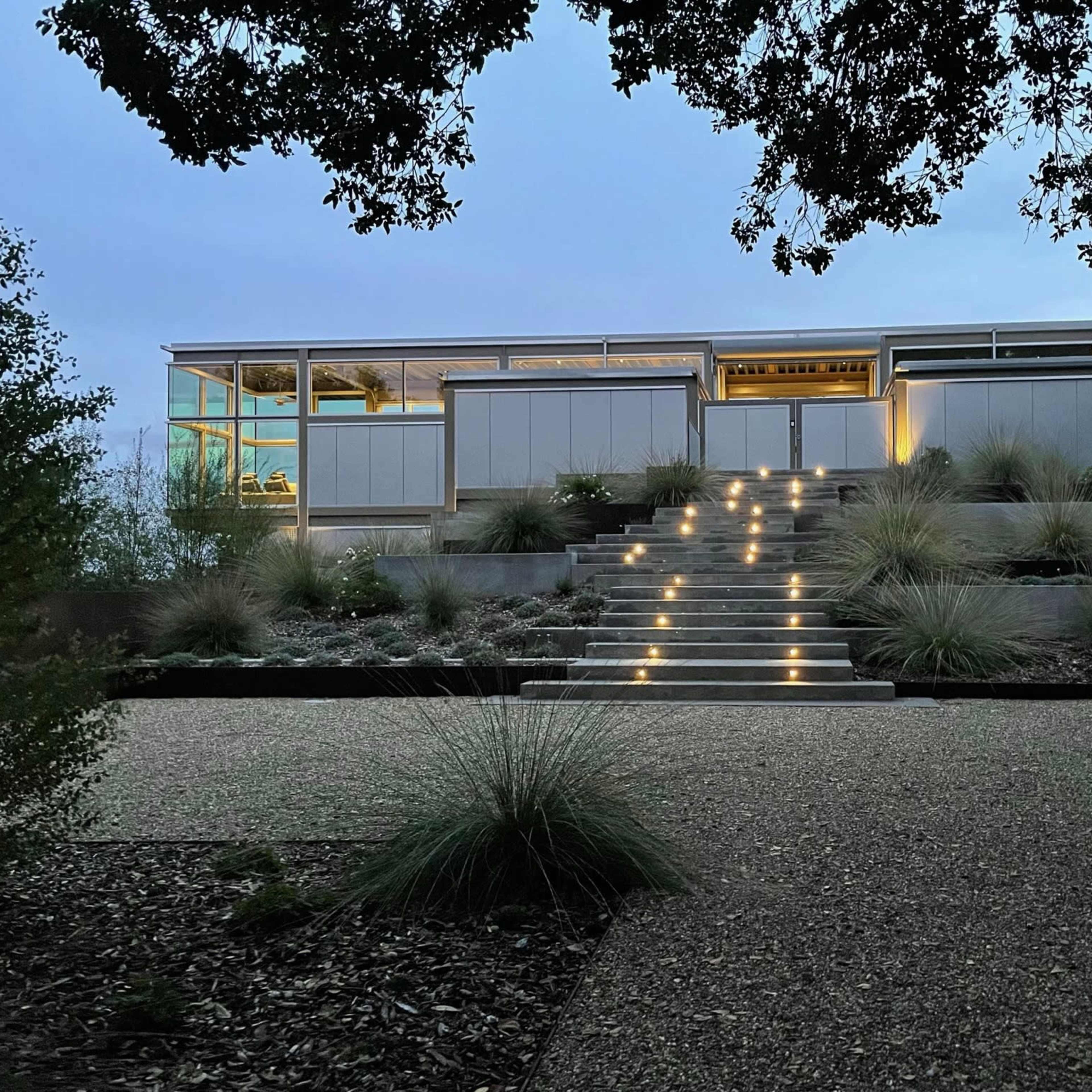 A modern house is illuminated by soft outdoor lighting near a stepped pathway leading up from a gravel driveway, surrounded by landscaped vegetation.