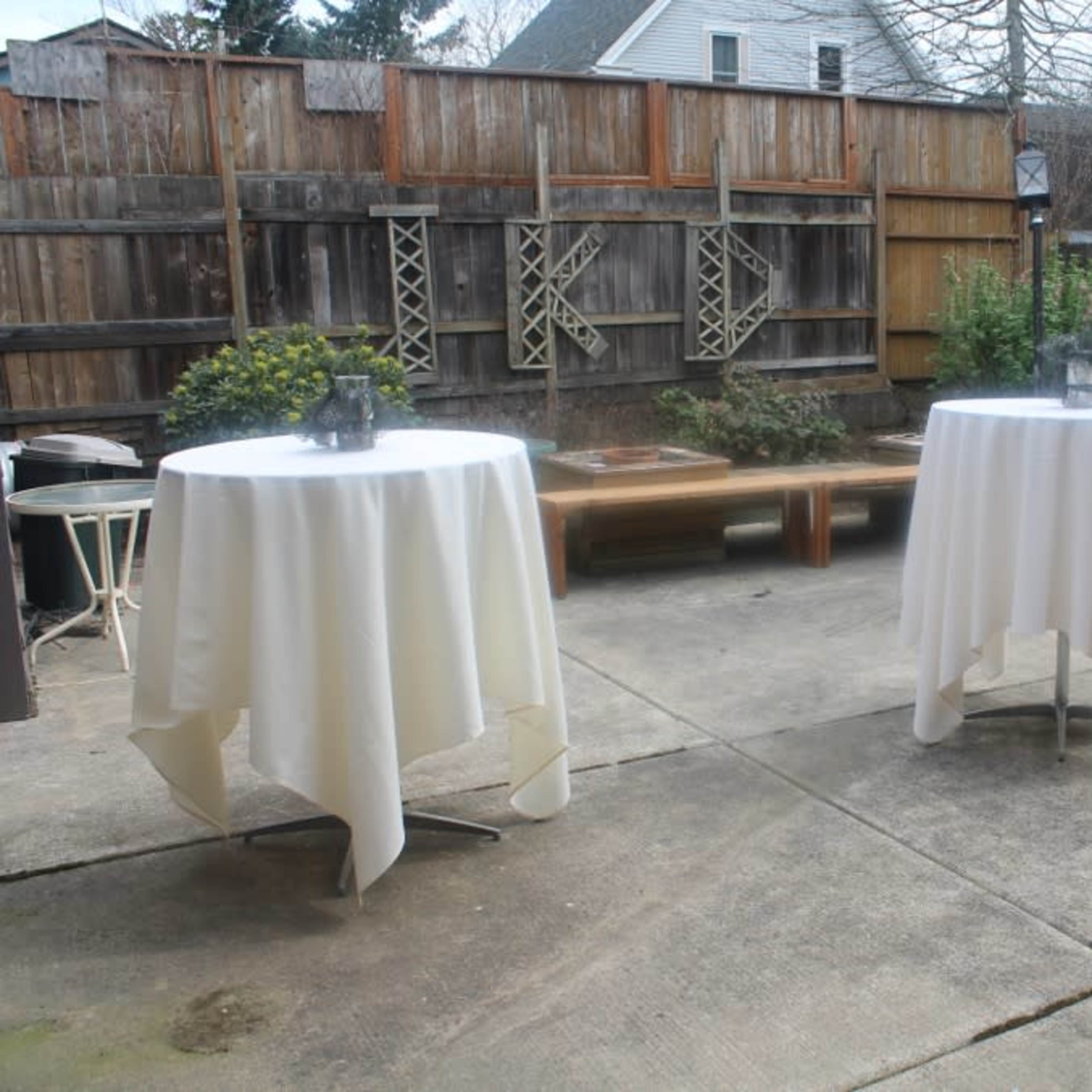 A patio with two round tables covered in white tablecloths, surrounded by a wooden fence and greenery.