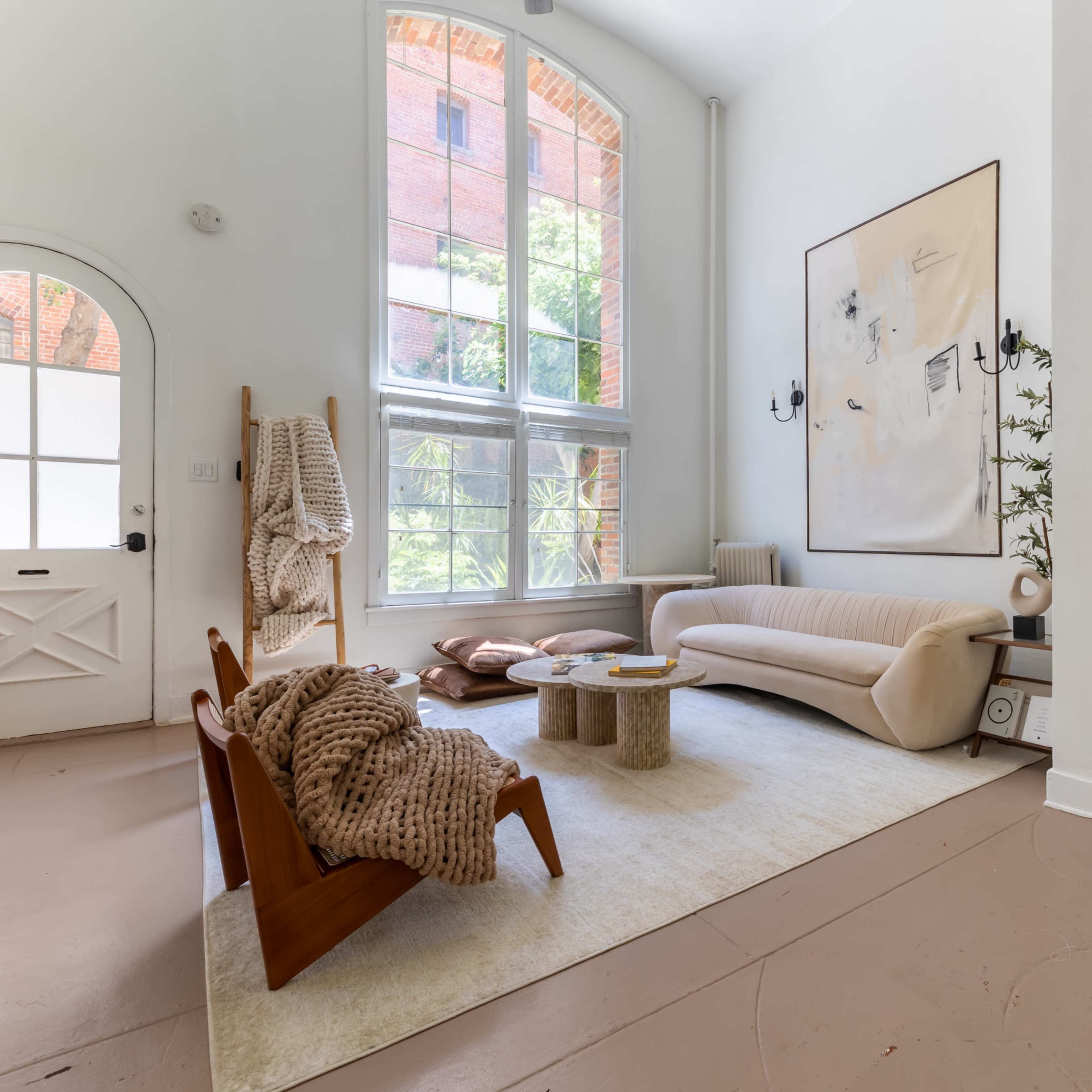 A bright, modern living room featuring a curved sofa, a stone coffee table, and a workspace in the corner, with large windows allowing natural light to fill the space.