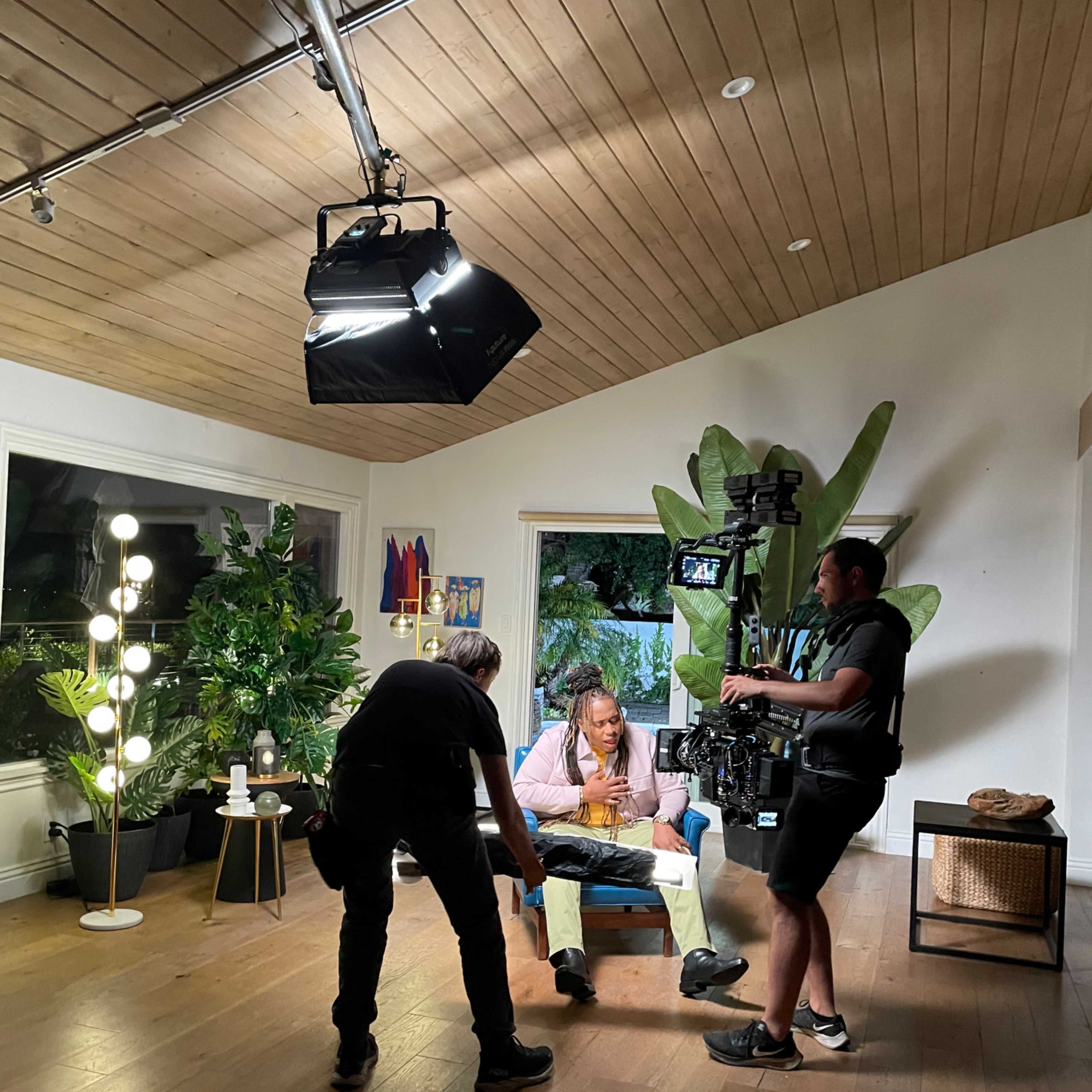 A film crew is setting up a shot in a brightly lit living room with large plants and artwork on the walls.