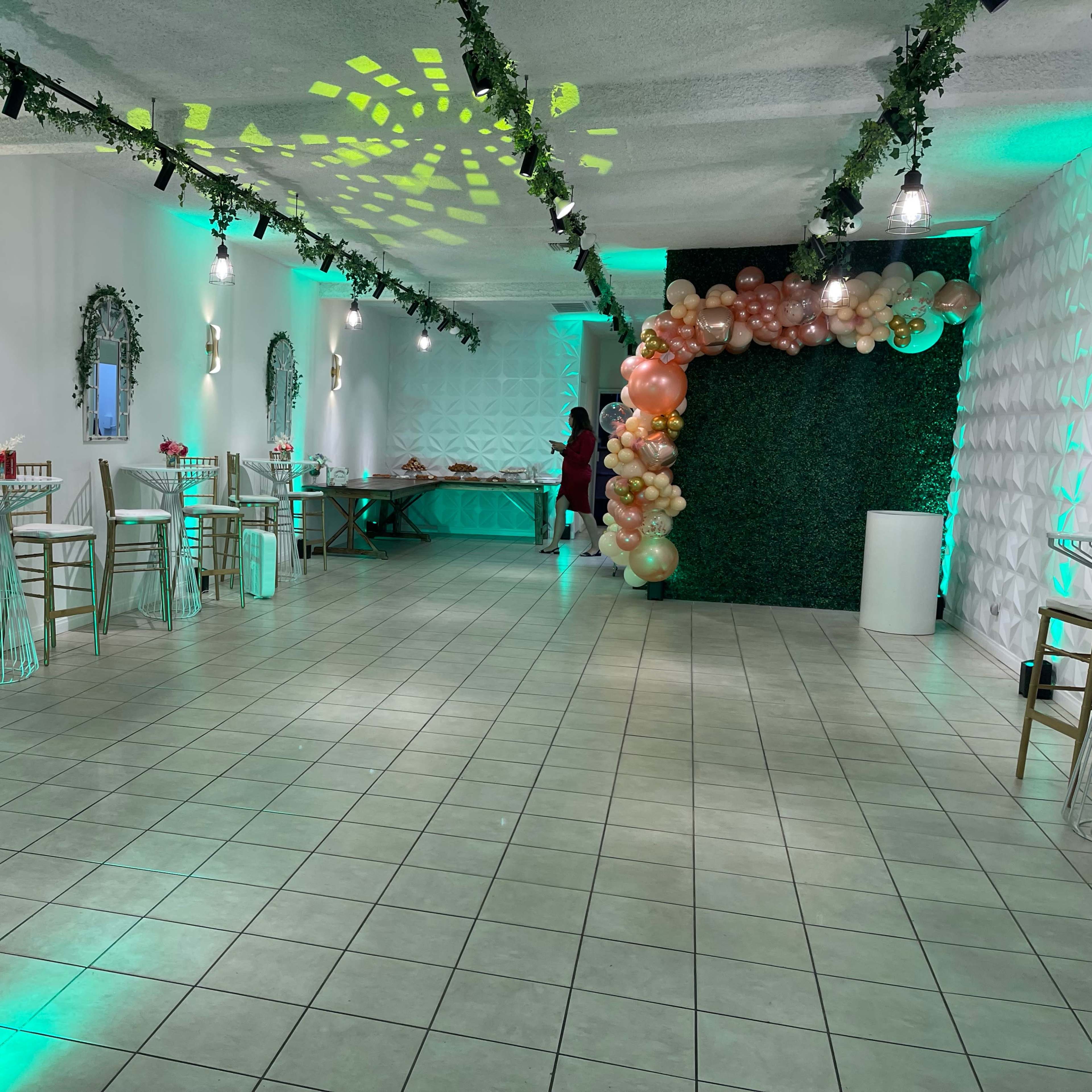 An indoor event space decorated with green lighting, tables, and a balloon arch in one corner.