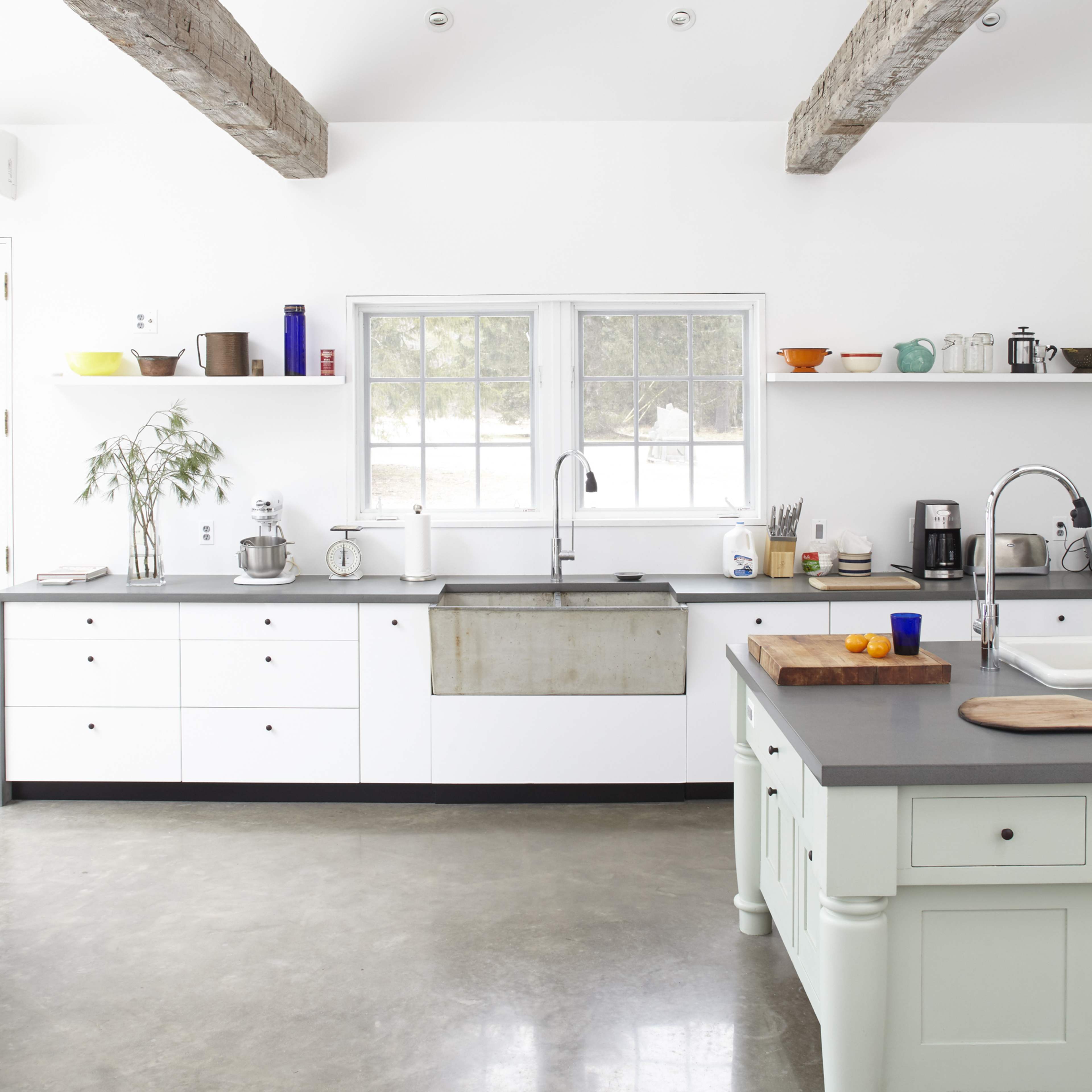 The kitchen features white cabinetry, a farmhouse sink, and exposed wooden beams above, with a large window allowing natural light to enter.