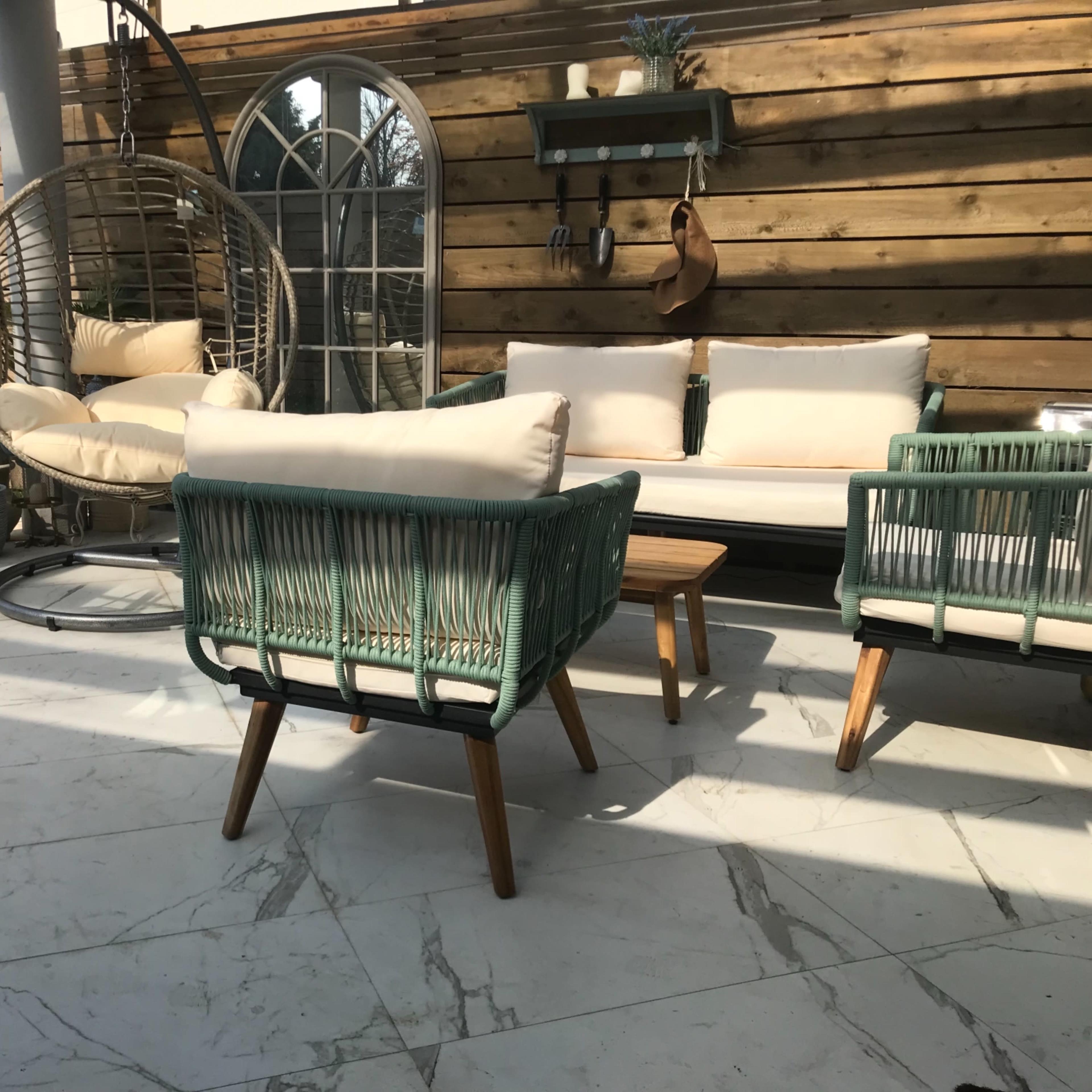 A patio area with a hanging chair, two green armchairs, a small wooden table, and a wooden wall in the background.